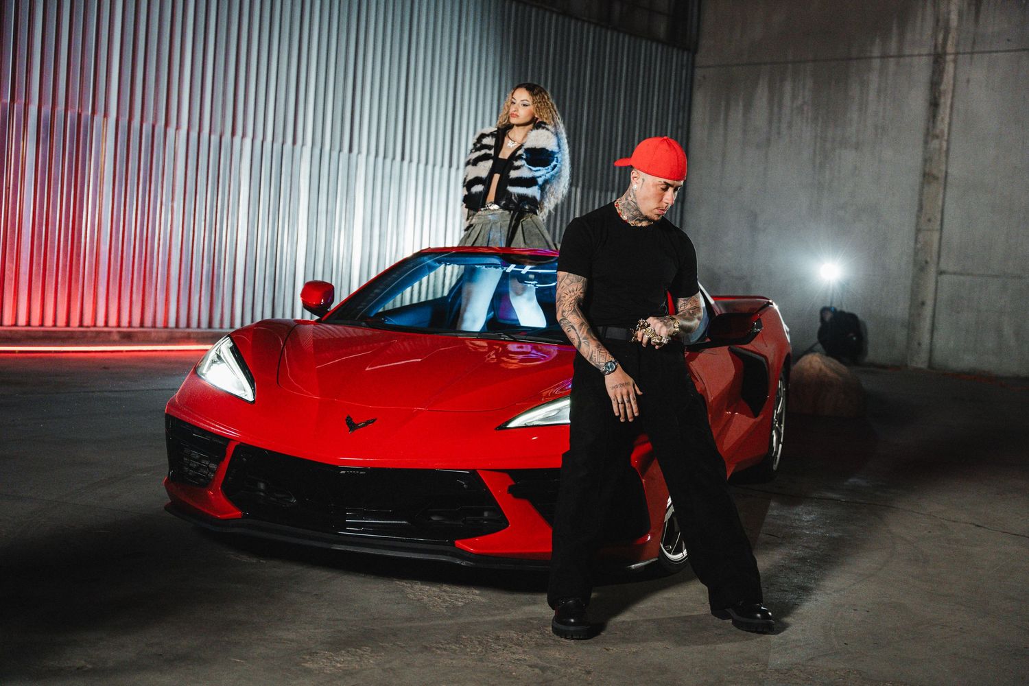 Fashion models pose with red sports car in industrial garage setting.