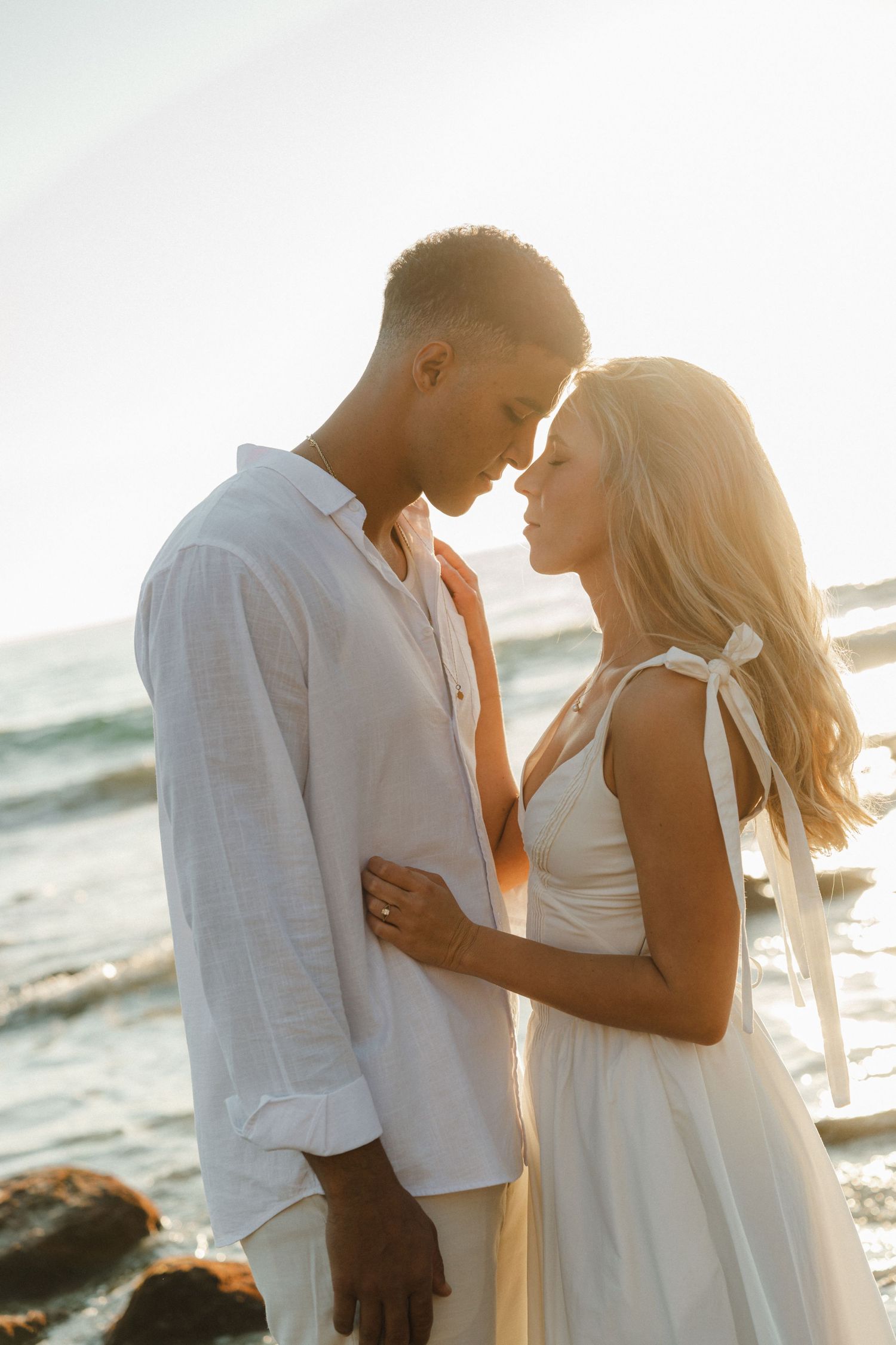 Romantic couple in white outfits embracing on a beach during golden hour sunset.