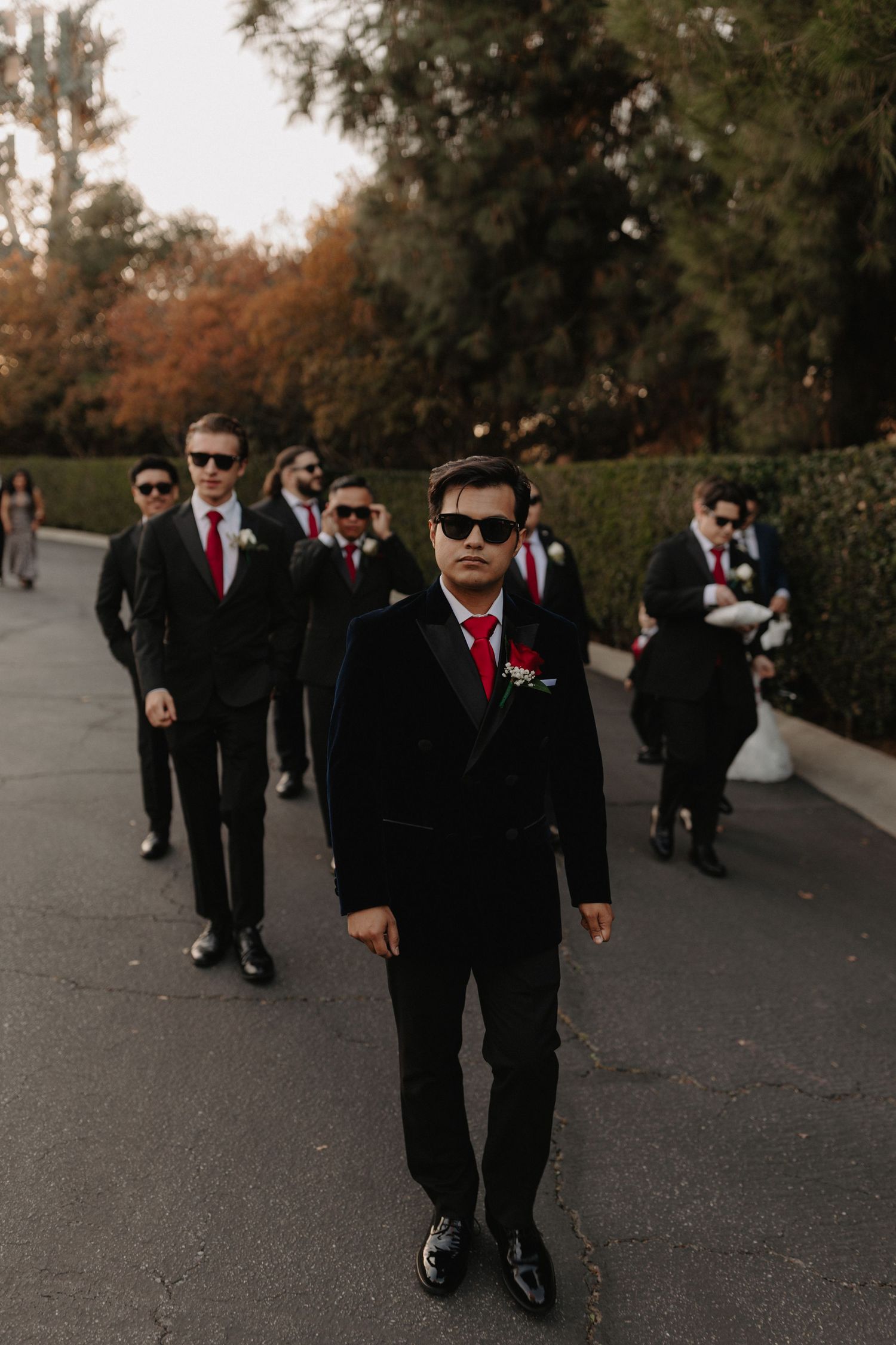 Groomsmen in black suits and sunglasses walk down a pathway in a stylish wedding party procession.