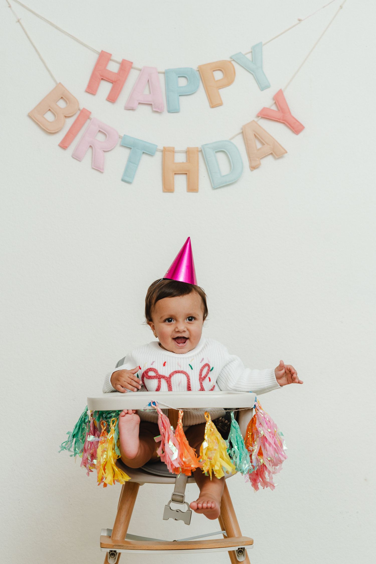 Baby celebrates first birthday in decorated high chair with pastel birthday banner and party hat.