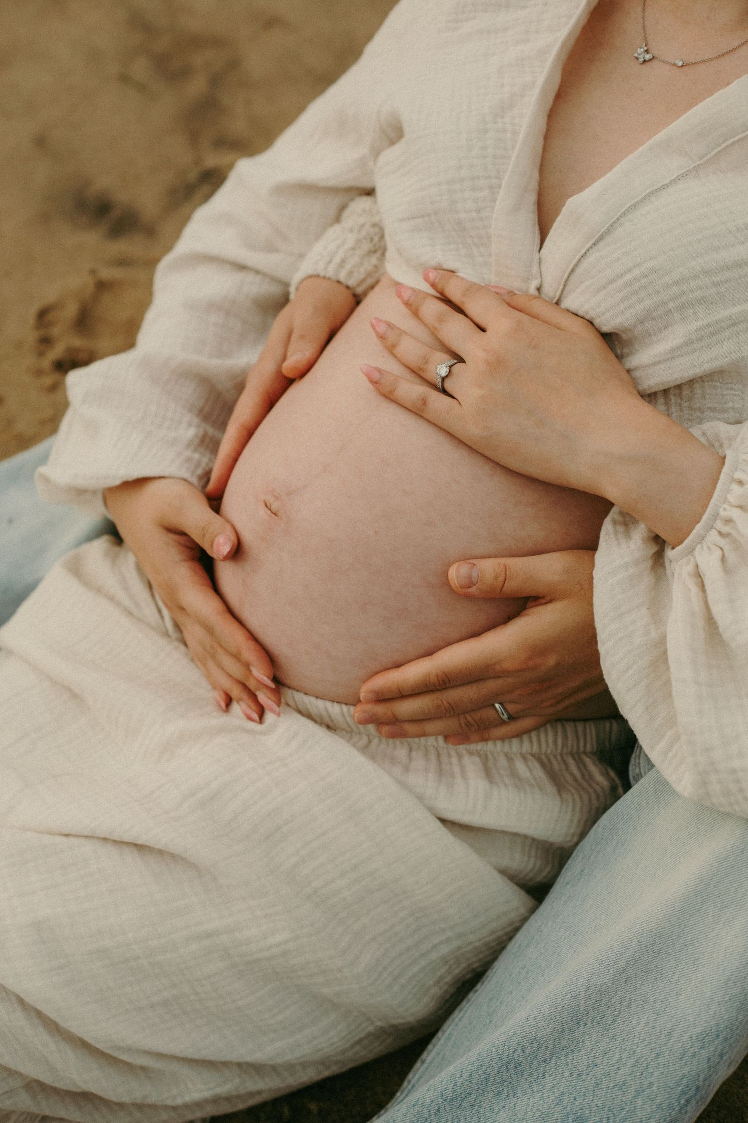 Pregnant woman in white linen dress cradling bare baby bump on beach, wearing ring and necklace.
