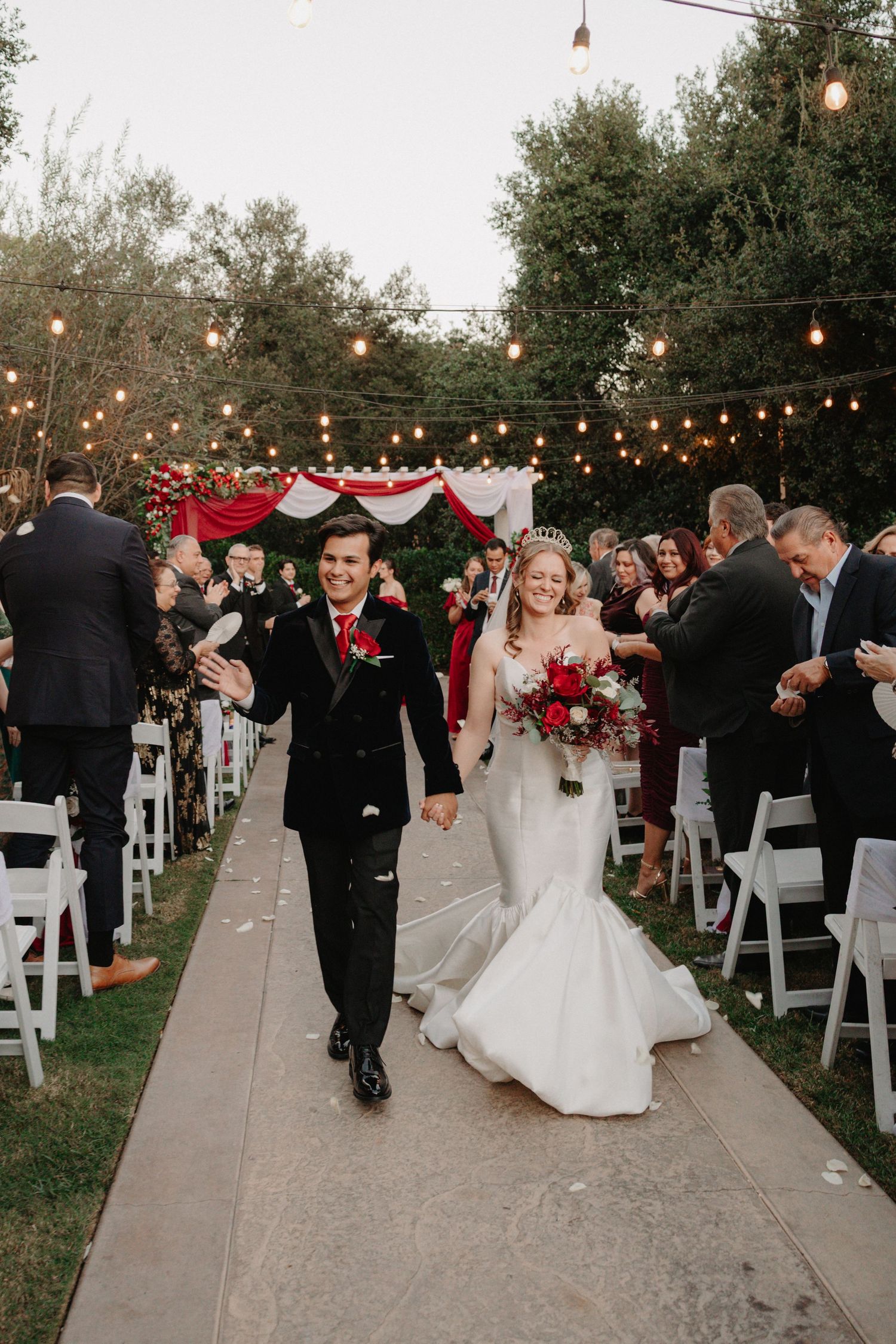Outdoor wedding ceremony at dusk with string lights illuminating guests and couple walking down aisle in garden setting.