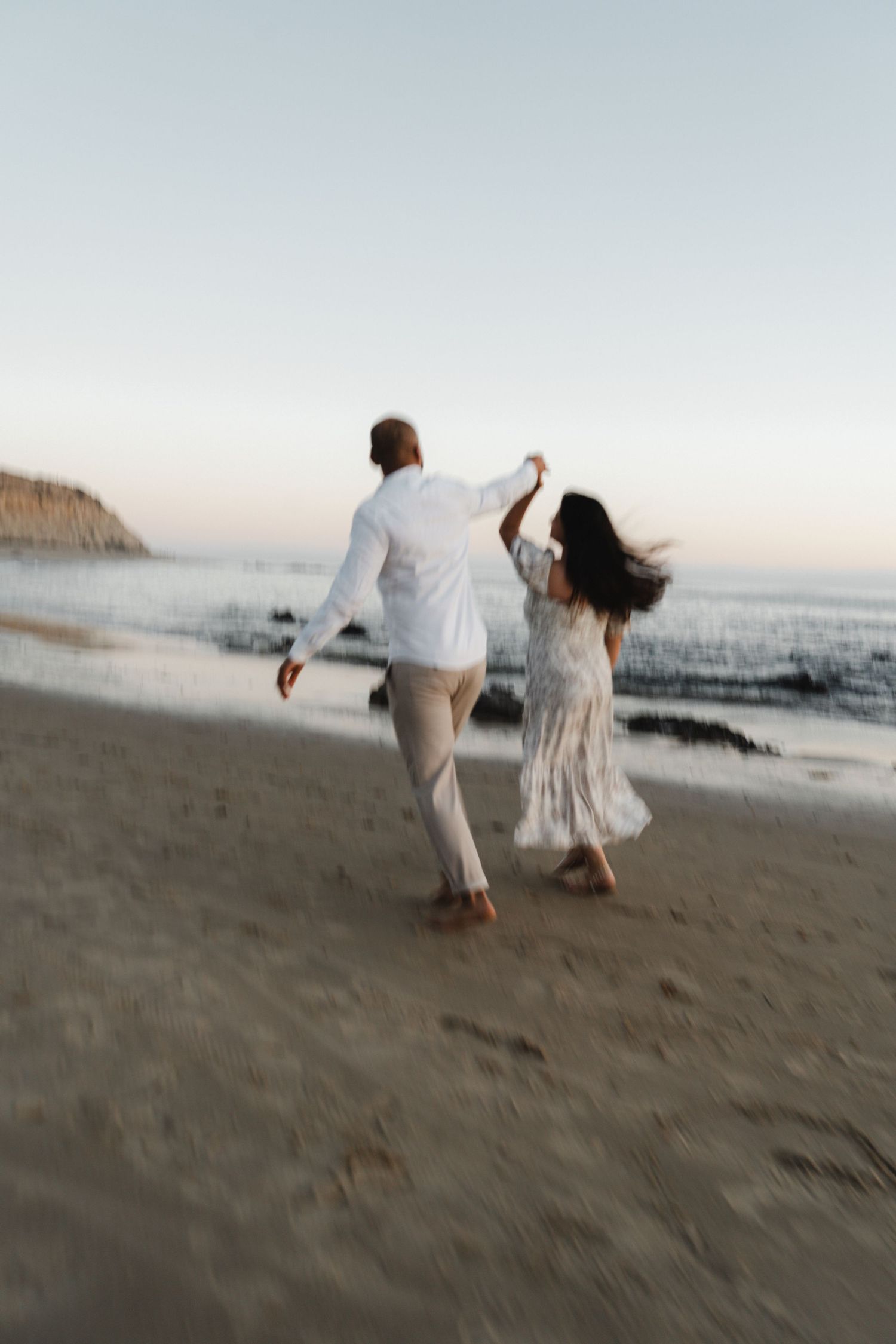 A couple in white outfits dances playfully together on a beach at sunset with waves and cliffs in the background.