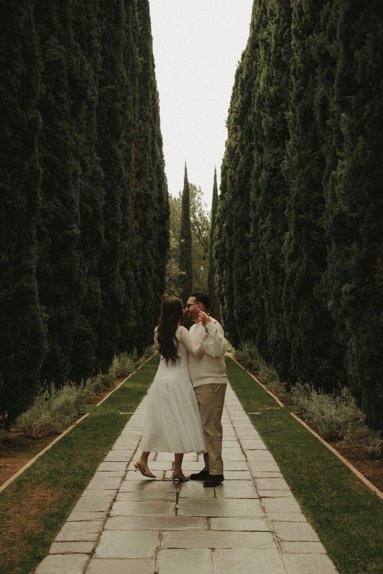 A couple in wedding attire walks down a dramatic cypress tree-lined path in a romantic garden setting.