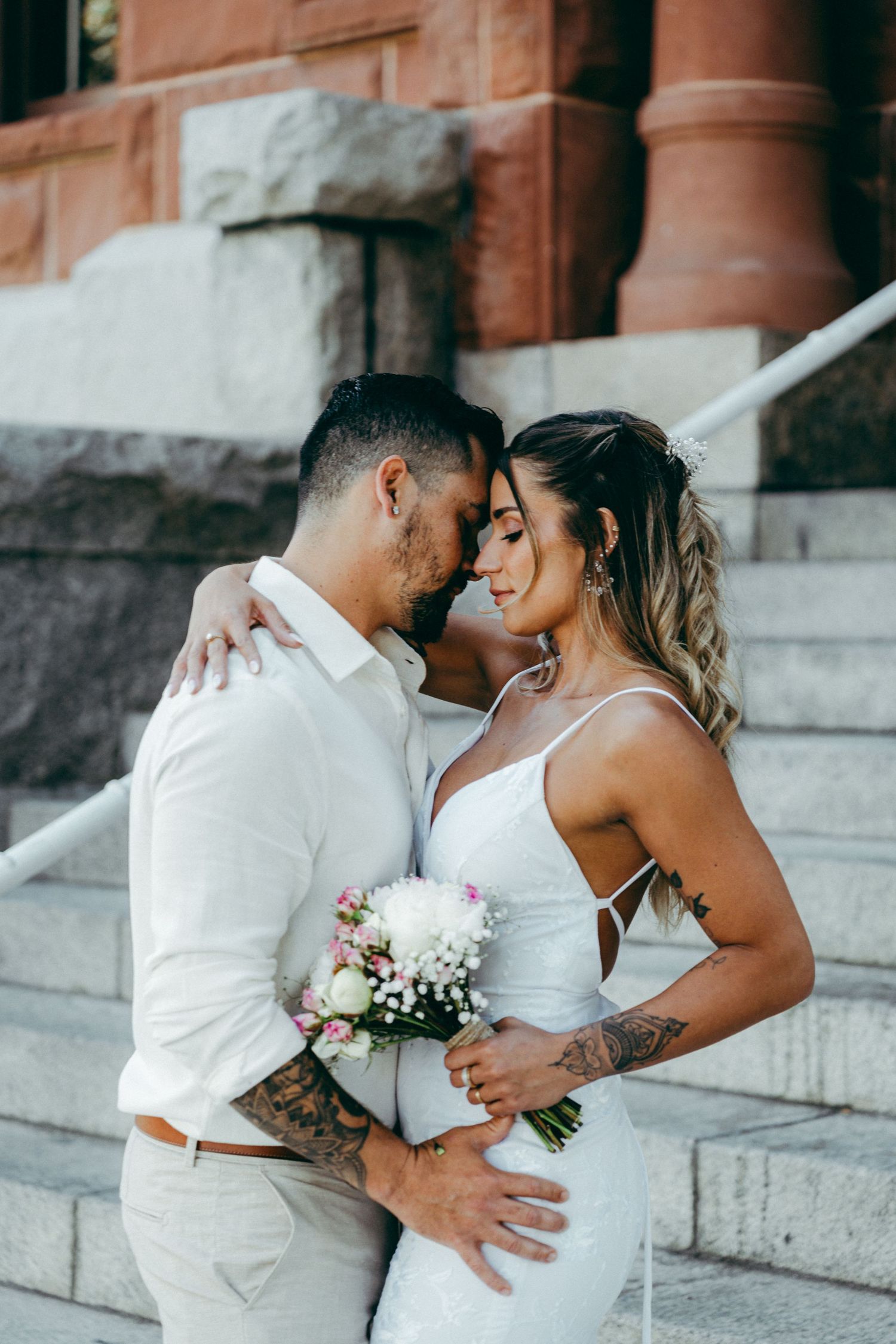A romantic wedding couple shares intimate kisses on stone steps while holding a delicate floral bouquet.