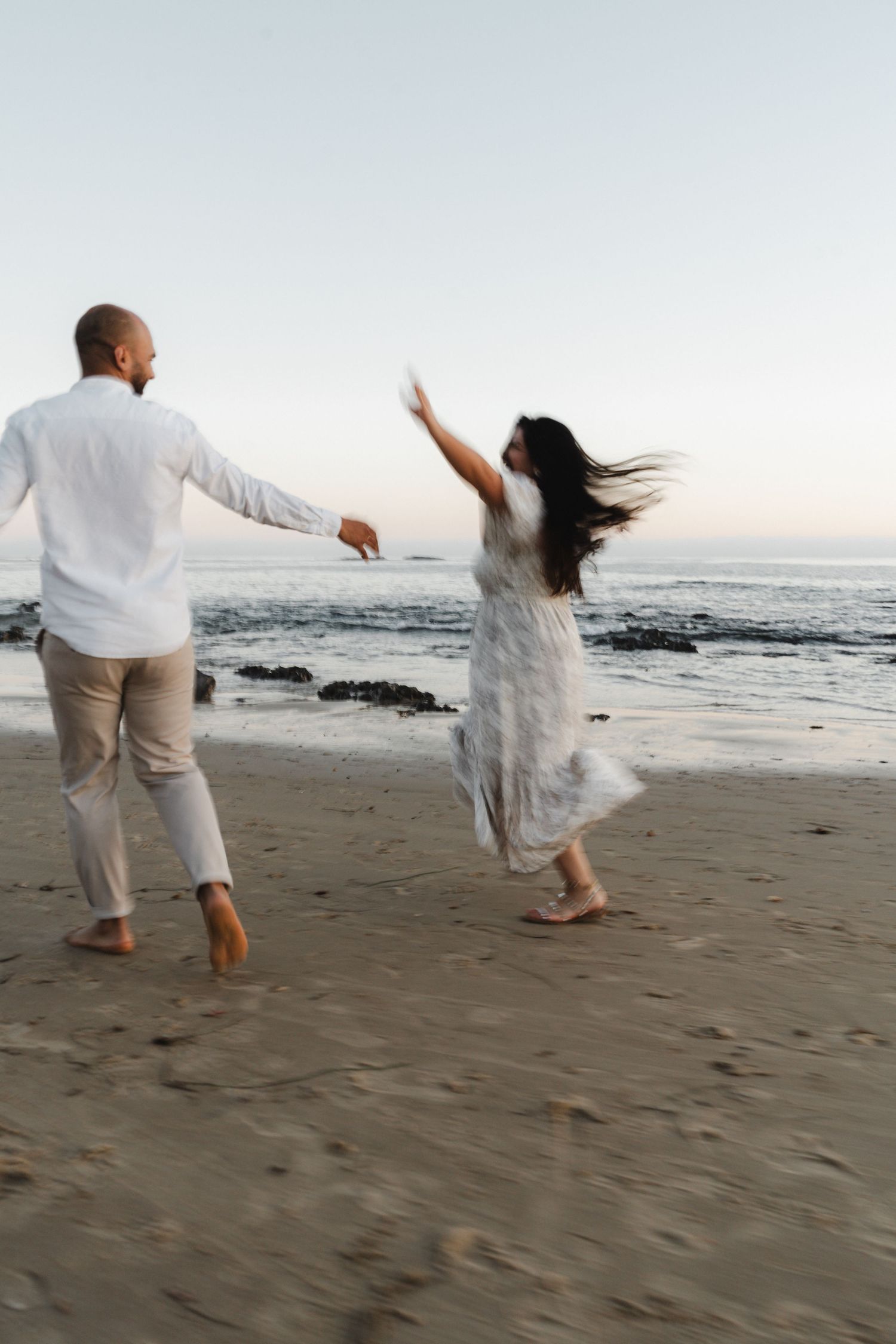 A couple in white clothing dances joyfully together on a sandy beach at sunset by the ocean.