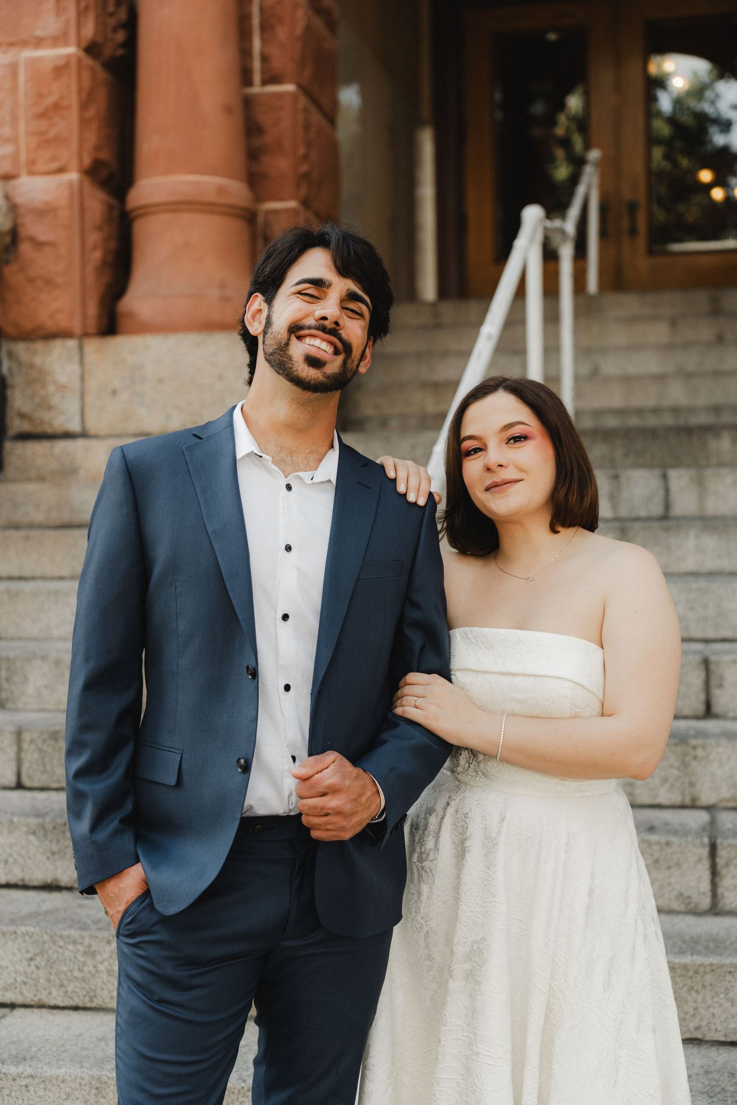 Wedding couple posing on stone steps in formal attire outside a historic building.