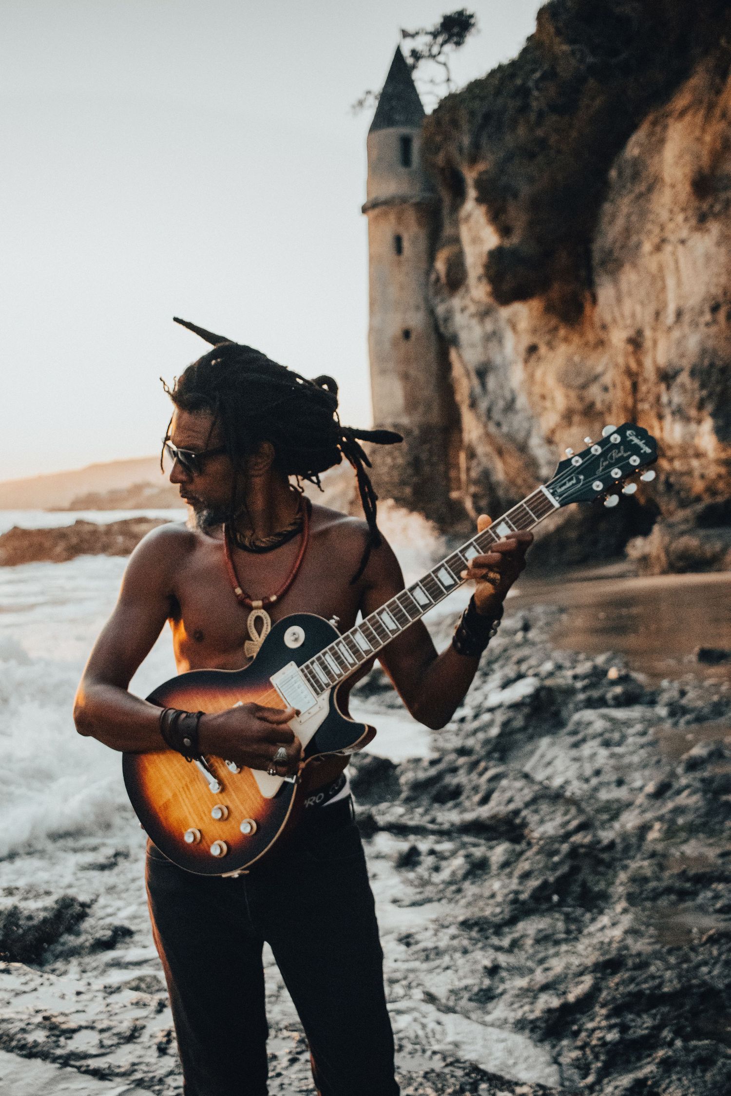 A musician plays guitar on a rocky beach coastline with cliffs and ocean waves in the background.