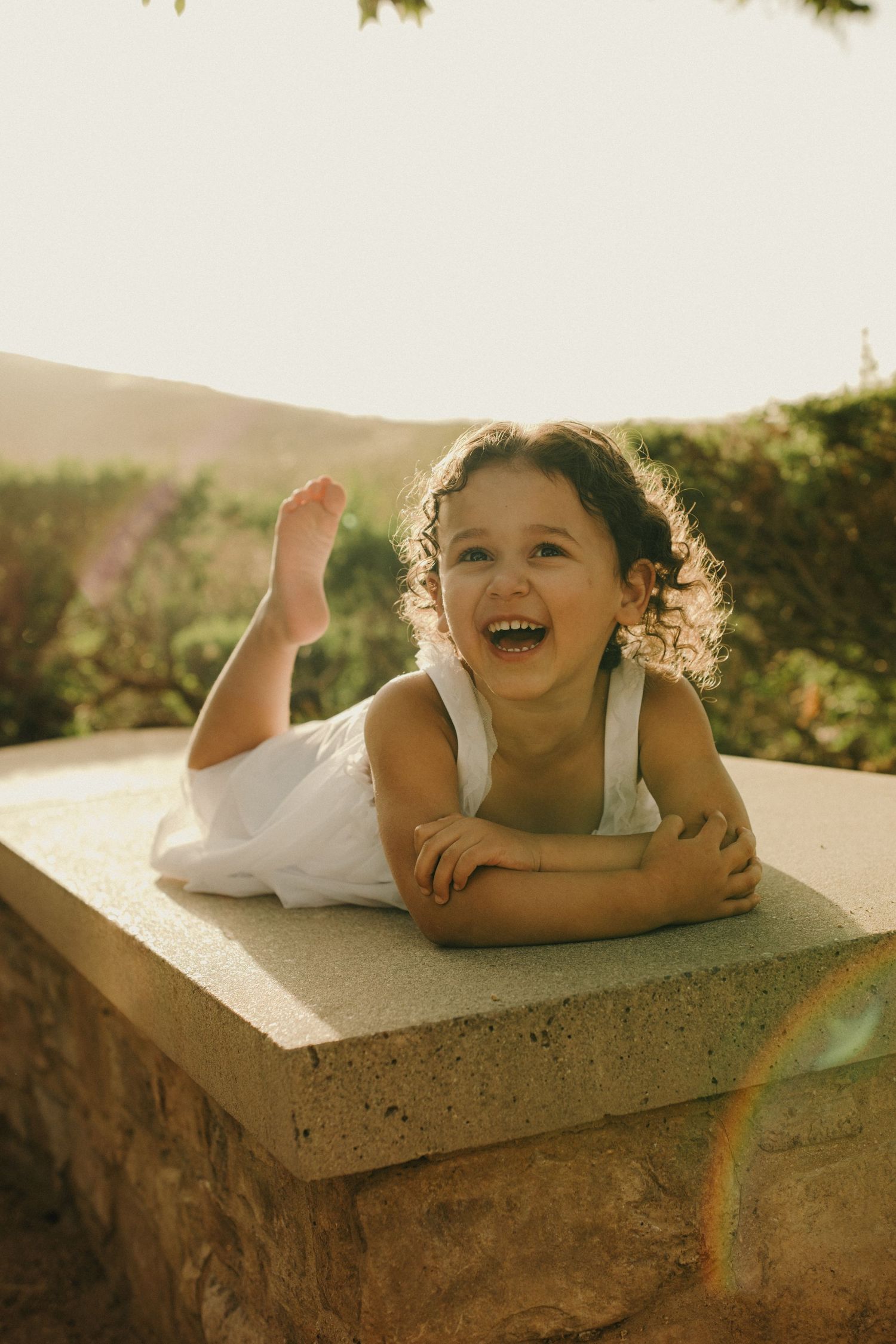 A joyful young girl in a white dress laughs while lying on a stone wall outdoors in warm golden sunlight.