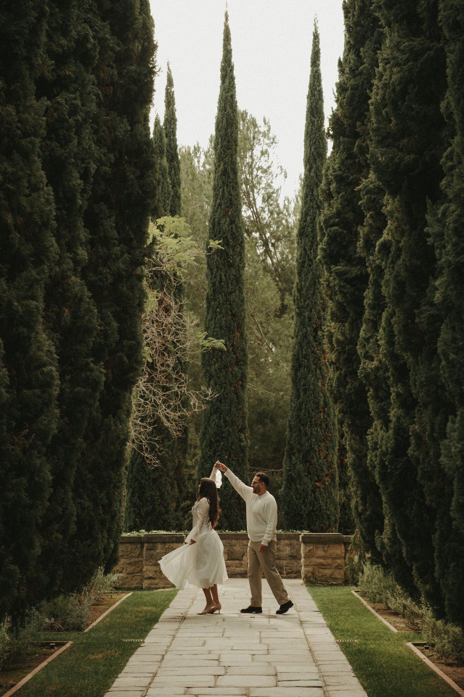 A couple dances joyfully on a stone path surrounded by towering cypress trees in a garden setting.