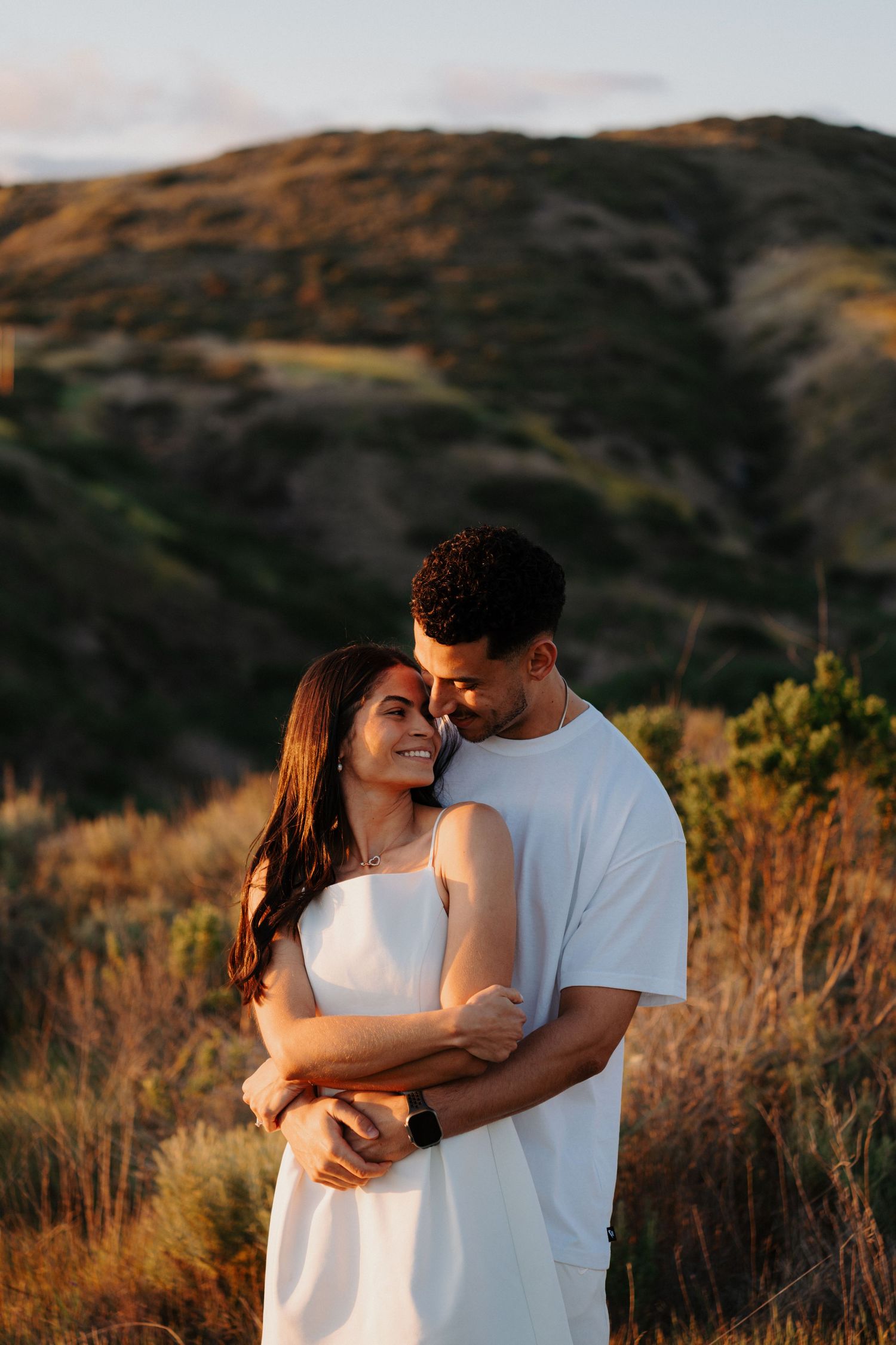 A couple shares intimate moments at sunset in a golden hillside setting wearing white outfits.