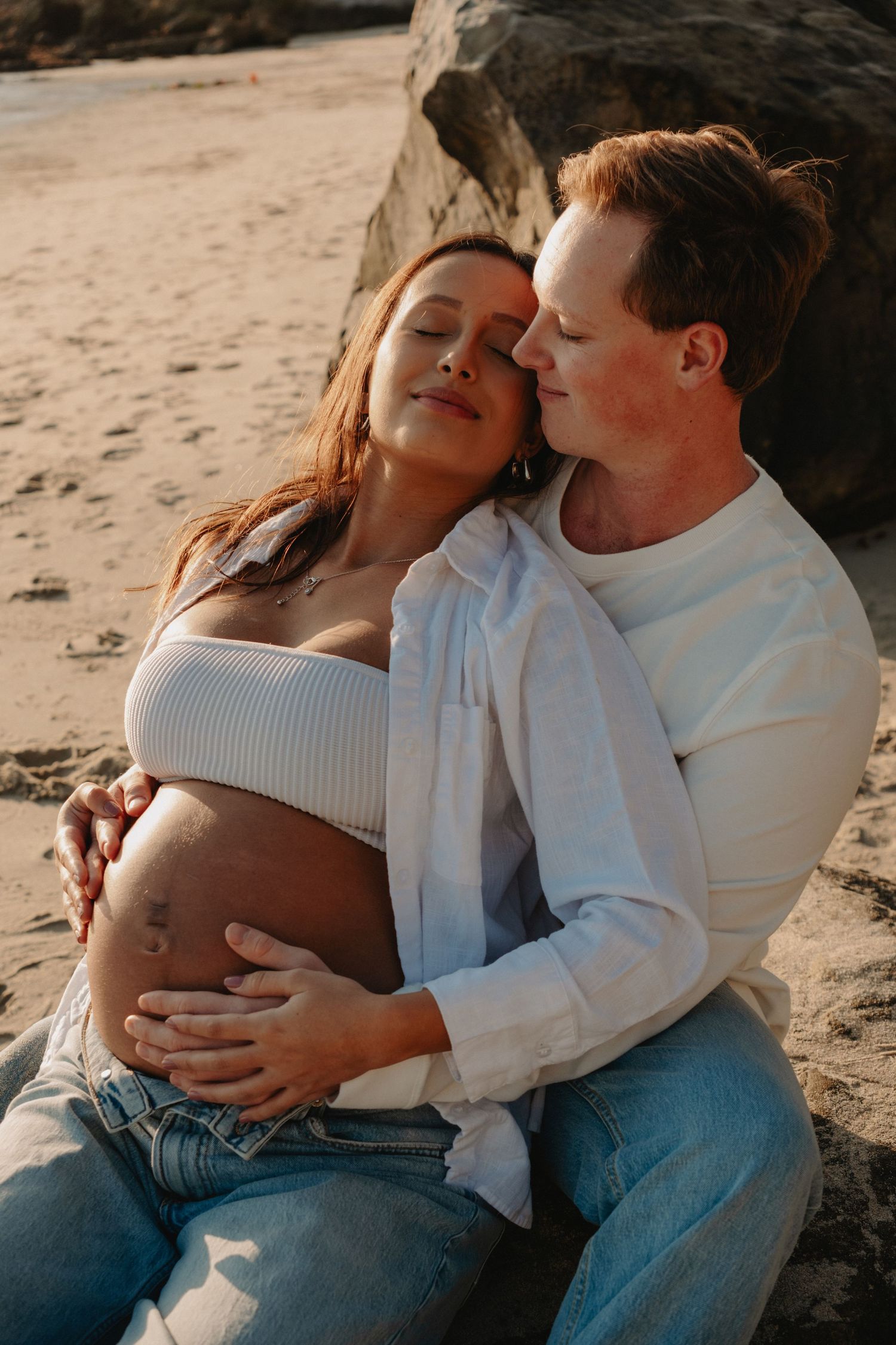 Intimate maternity photoshoot on a beach showing a couple in white and denim embracing.
