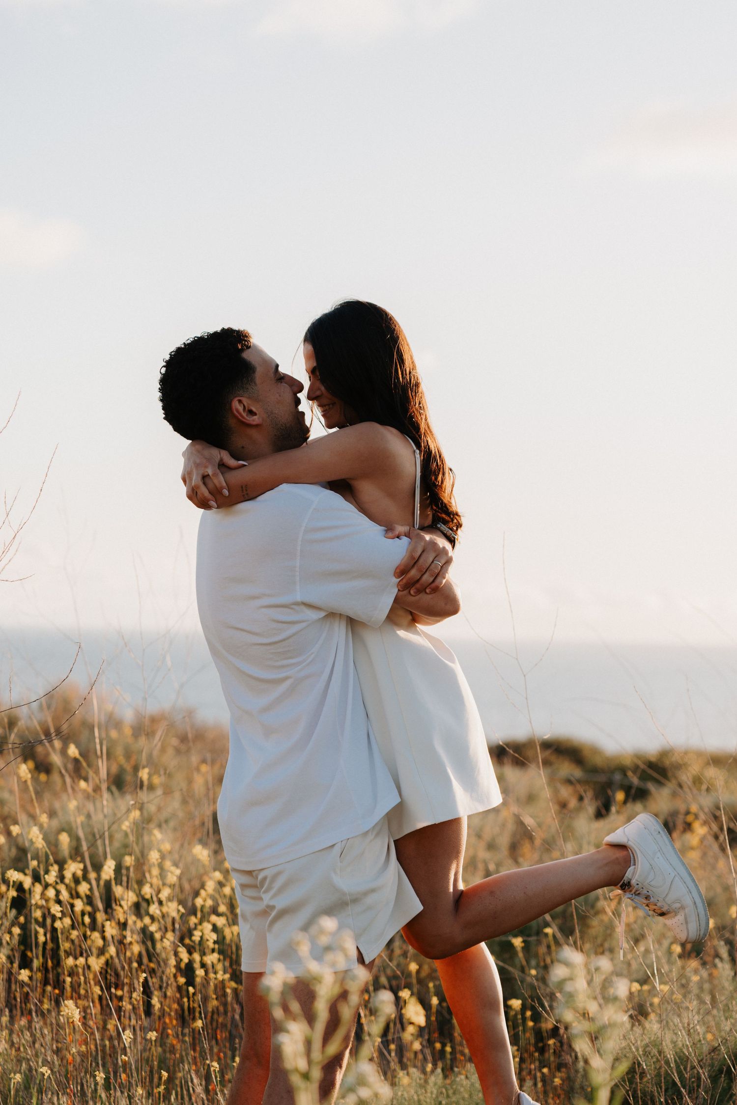 A romantic couple in white outfits embrace and twirl together in a sunlit meadow with dried wildflowers at sunset.