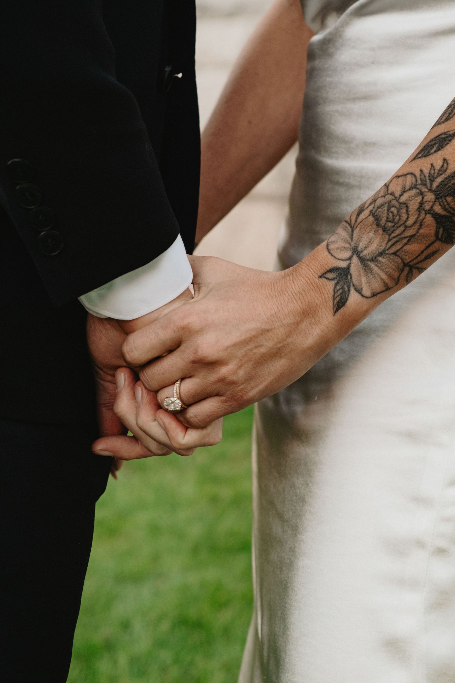 Close-up view of newlyweds holding hands with visible engagement ring and floral tattoo against white dress and dark suit.