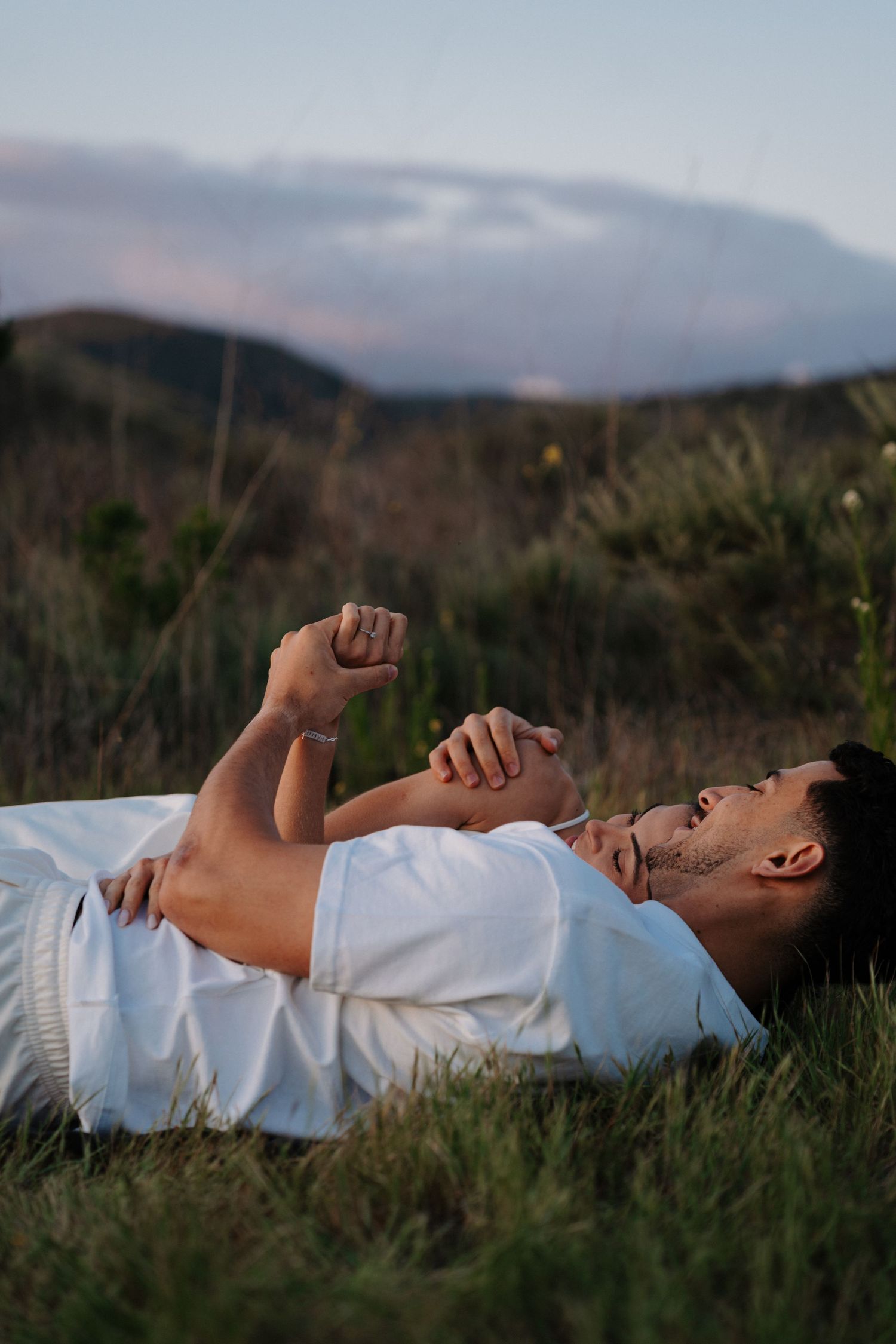 Person in white shirt lying in grass against mountainous landscape at dusk.
