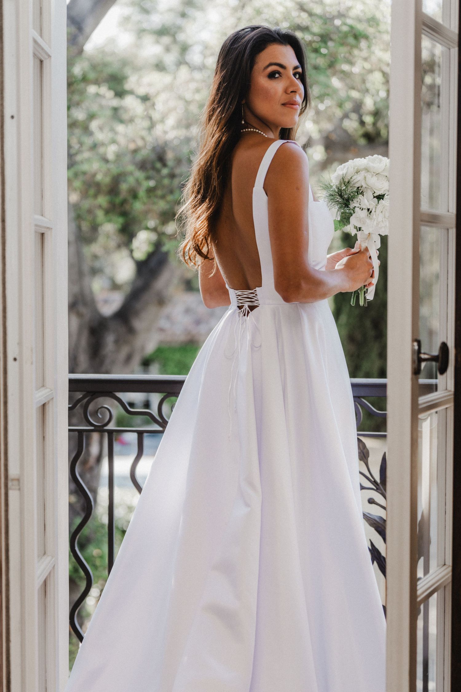 A person in a flowing white dress poses in front of French doors in an elegant setting.