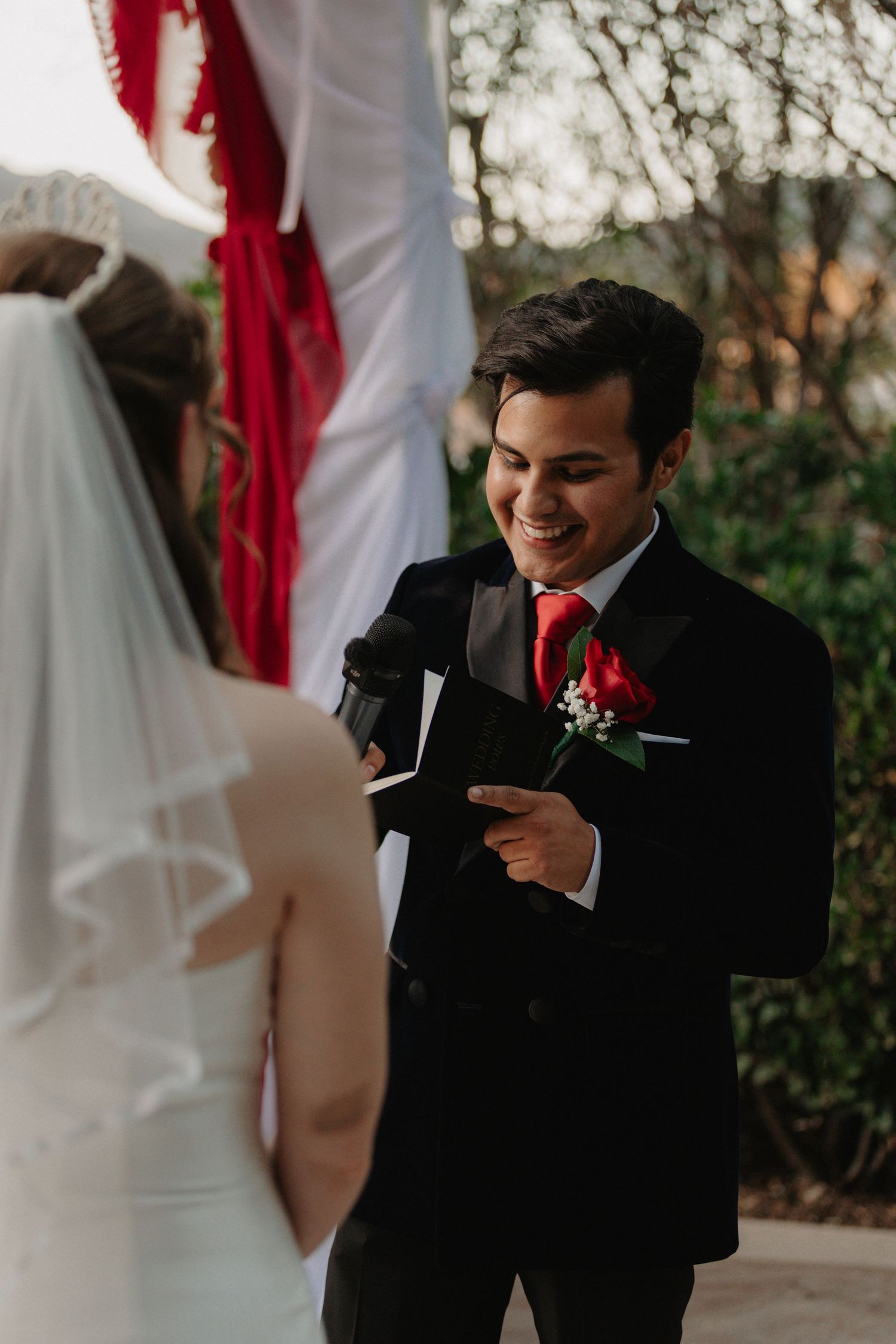 Groom in black tuxedo smiles while looking at phone during wedding ceremony.