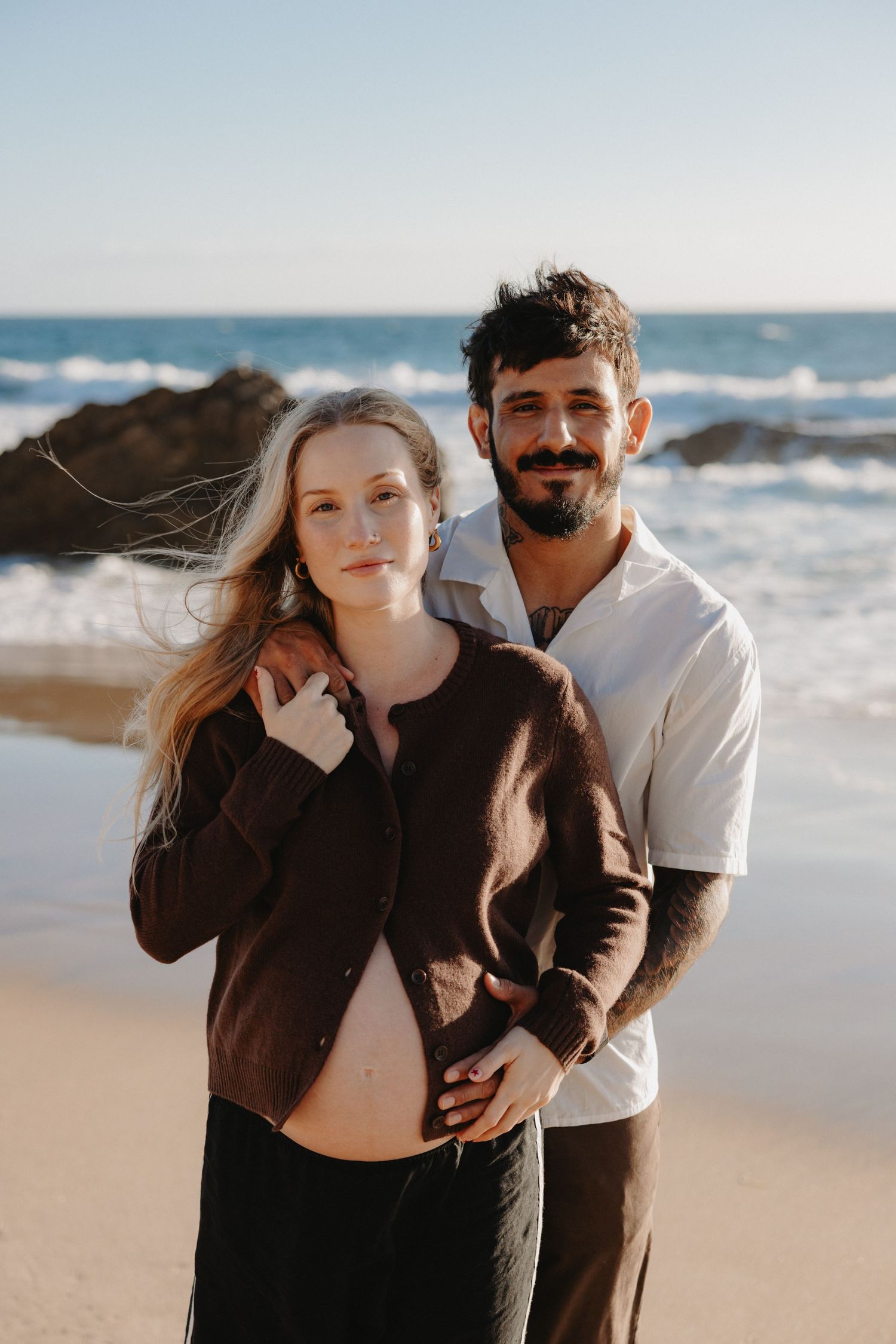Romantic couple embraces on a sandy beach with ocean waves and rocky coastline in the background.