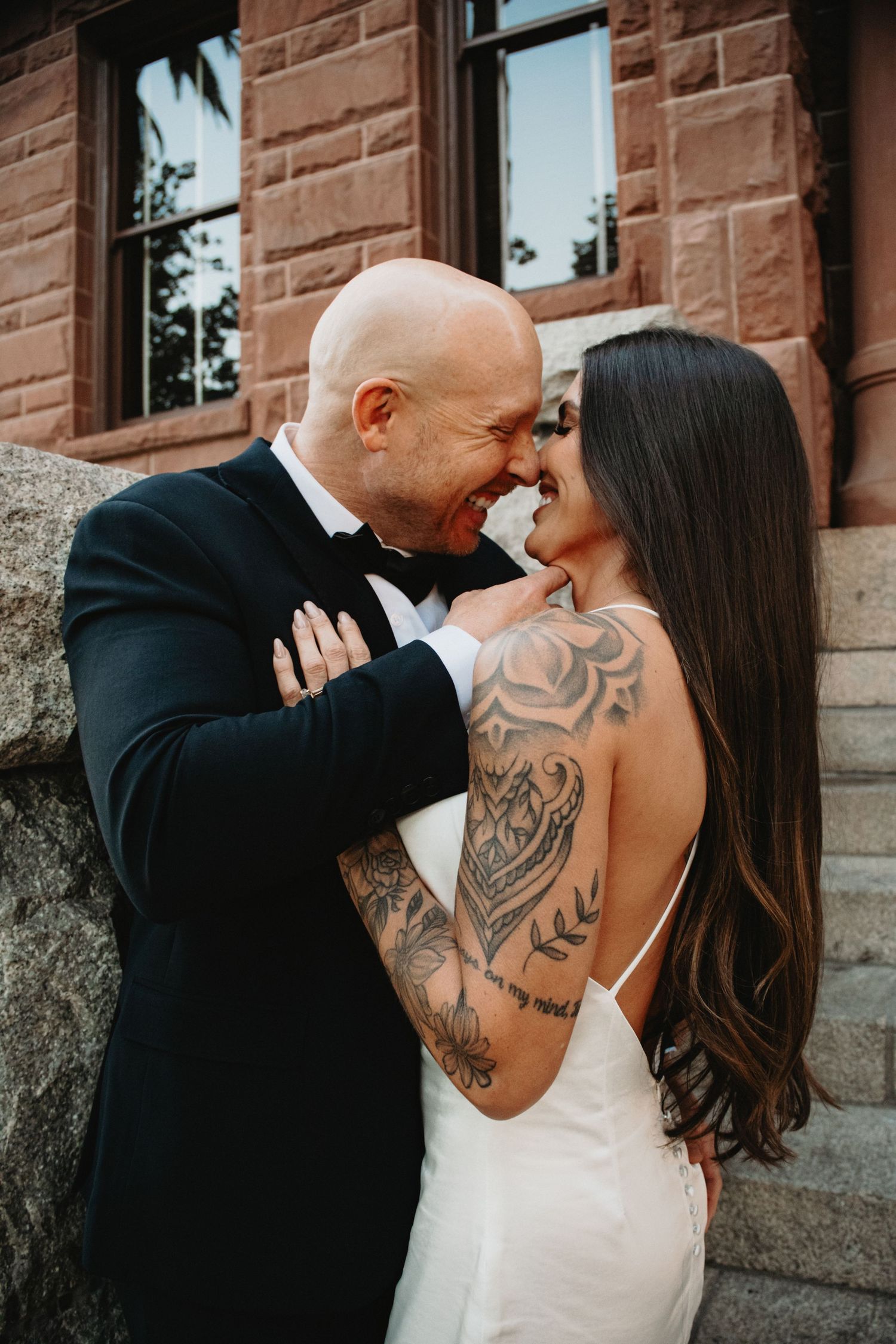 A romantic wedding couple embraces and kisses against a brick wall, with the bride showing elegant back tattoos.