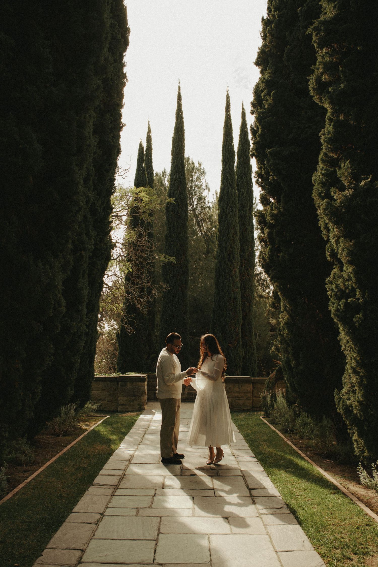 Two people walk along a stone path lined with tall cypress trees in a garden setting during golden hour.