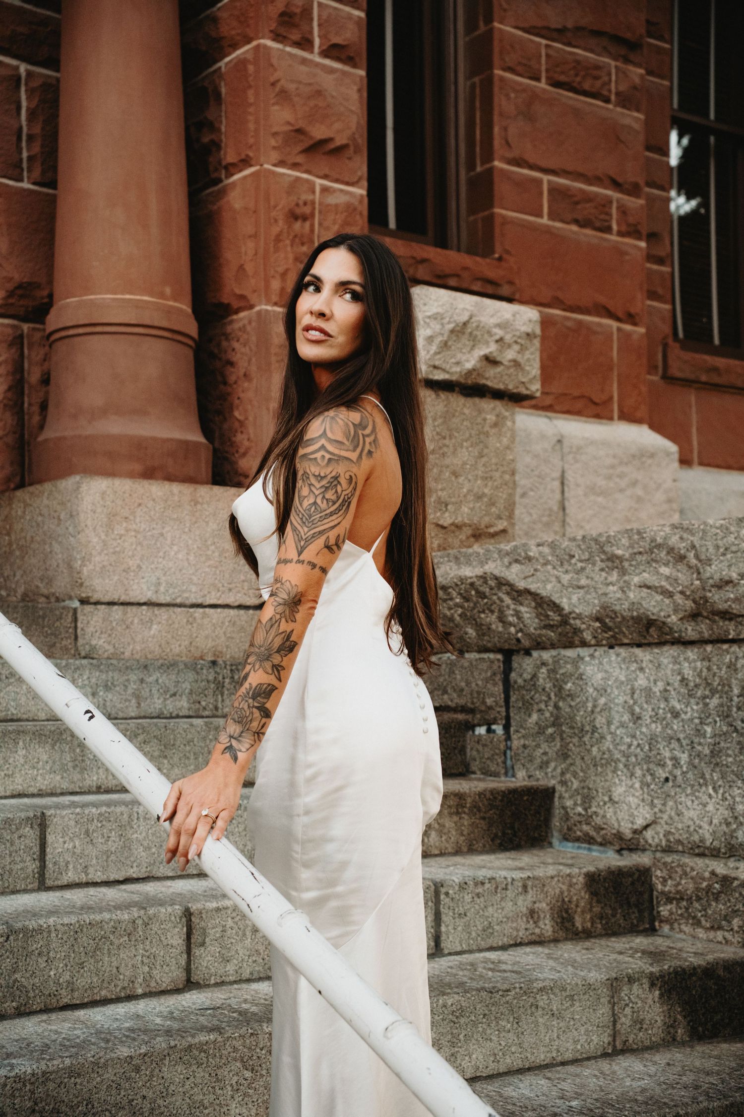 A woman in a white dress sits on stone steps near a brick building with columns, showing a tattoo on her arm.