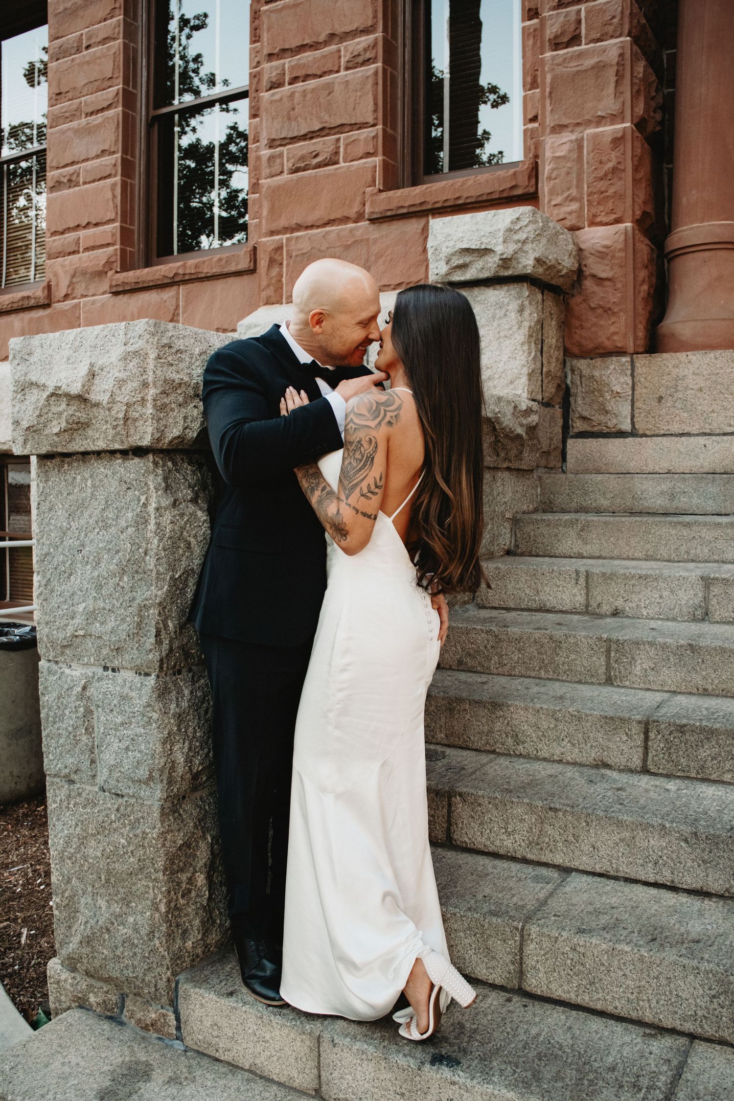 A romantic wedding couple shares a tender kiss against a rustic stone building wall.