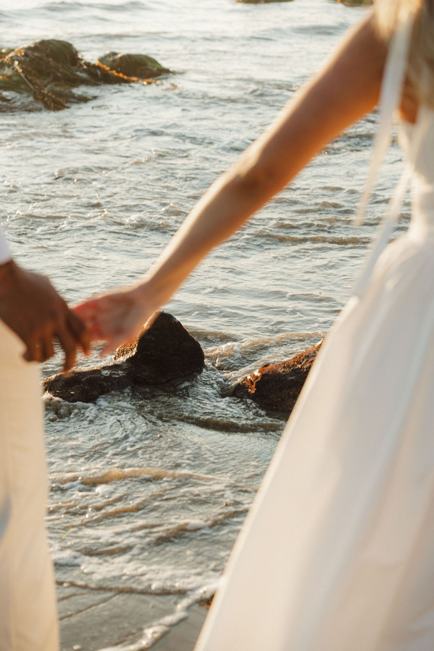 Two hands reaching towards rocks in shallow ocean water at sunset with white fabric visible in the foreground.