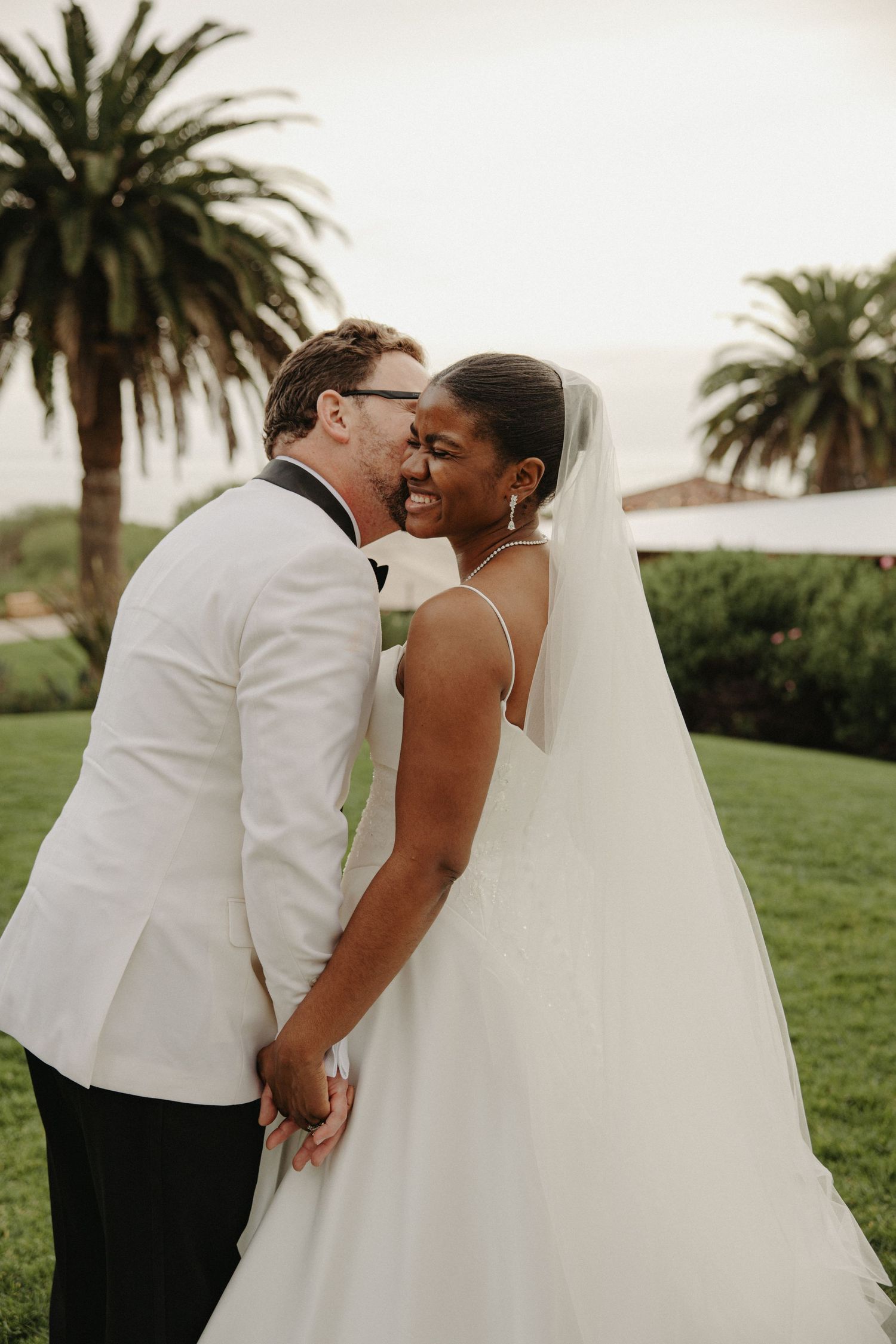 A bride in a white gown and veil smiles as her groom in a white tuxedo whispers in her ear outdoors.