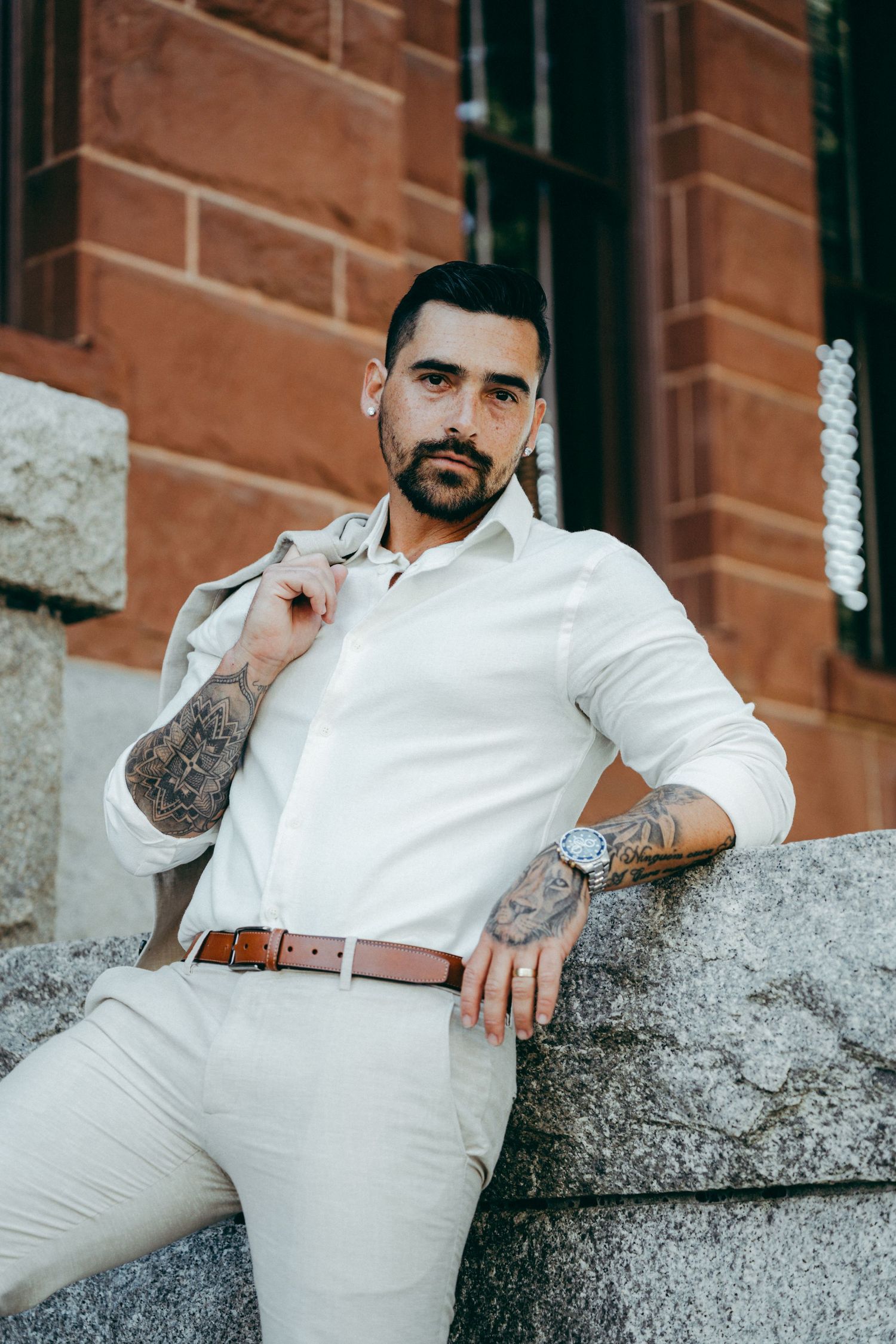 Man in white shirt and brown belt poses casually against brick wall with tattoos visible.