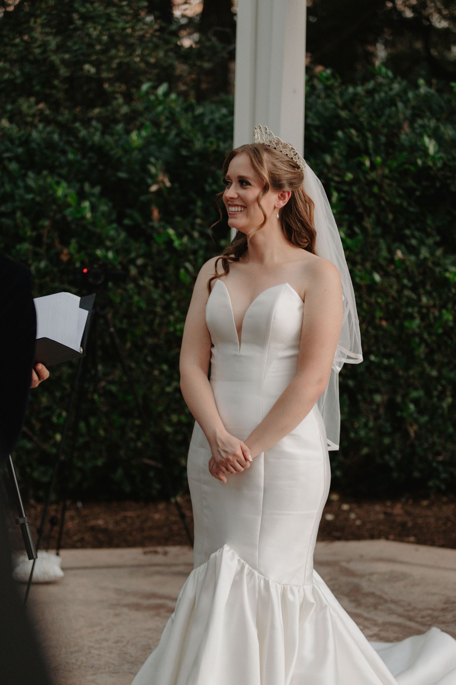 A bride in a stunning white mermaid wedding gown with sweetheart neckline and veil stands during outdoor ceremony.