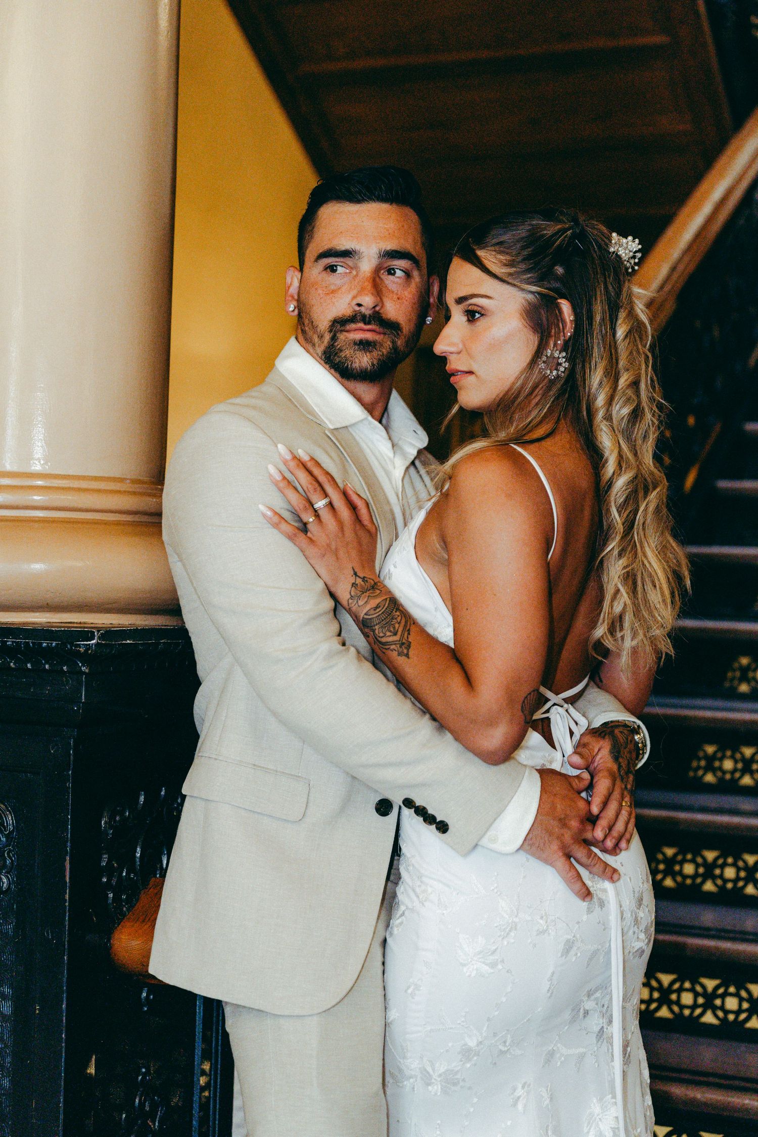 A couple in white wedding attire embraces near a staircase with warm lighting.