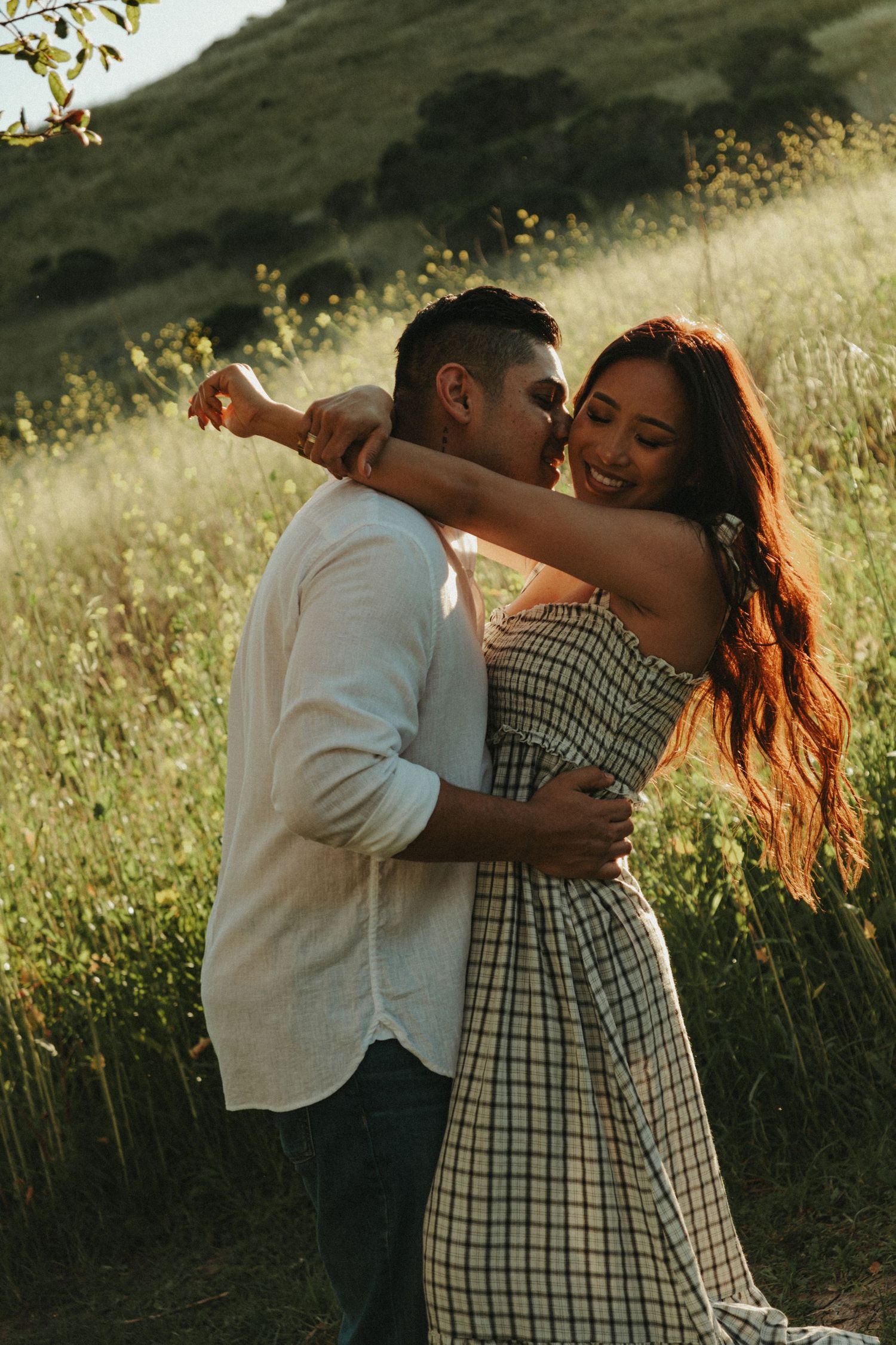 A couple embraces and dances in a sunlit meadow, the woman in a plaid dress smiling as her partner kisses her cheek.