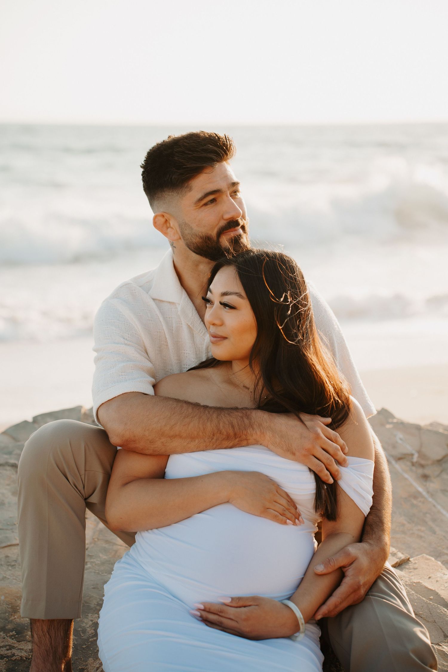 Romantic beach maternity photos with couple sitting together on rocky shore at sunset.