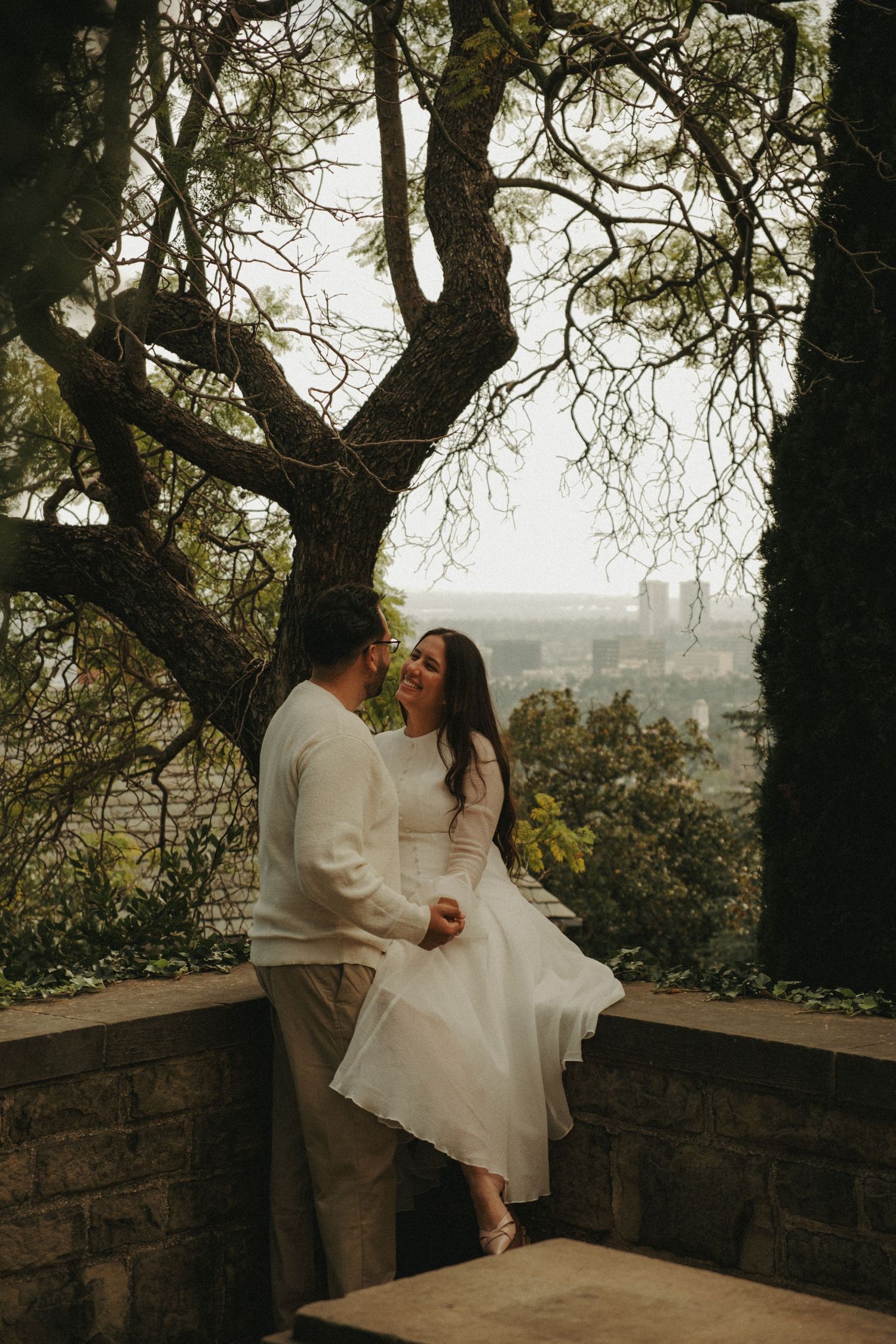 A romantic wedding photoshoot on a stone wall overlook with trees framing multiple intimate moments at sunset.