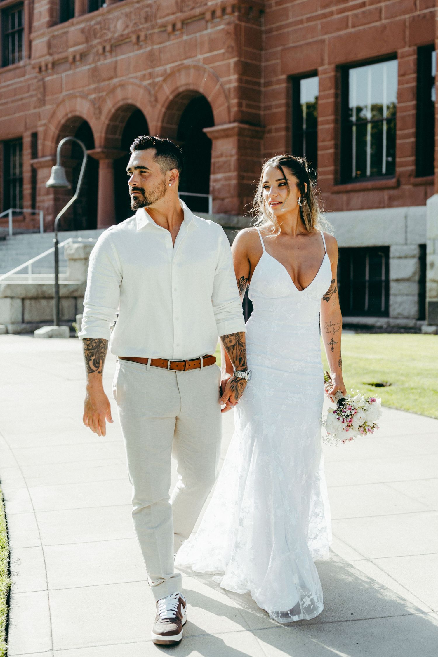 A couple in all-white wedding attire walks together outside a brick building.