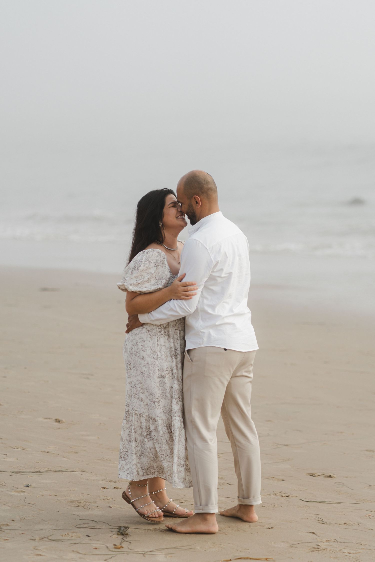 A couple shares a romantic embrace on a misty beach while wearing white and floral outfits.