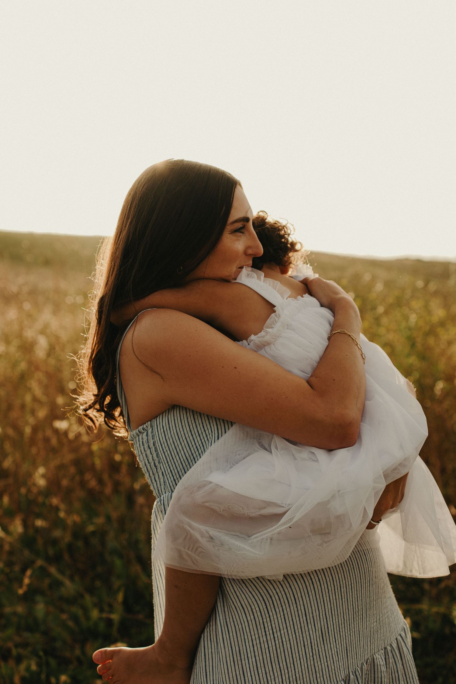 A mother tenderly holds her young child in a golden field at sunset, both dressed in soft, flowing outfits.