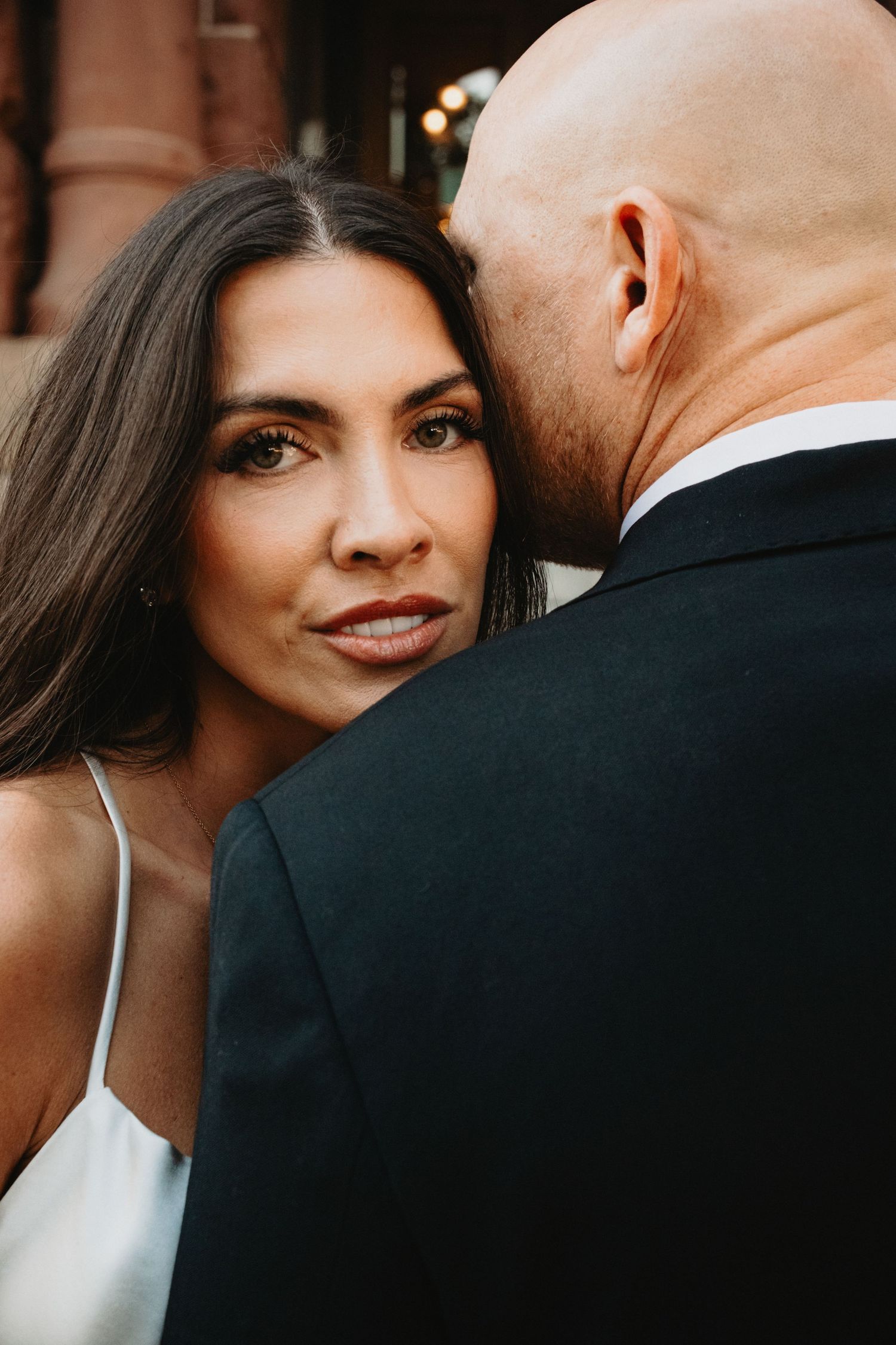 Close up portrait of bride with long dark hair in white dress standing next to groom in black suit.