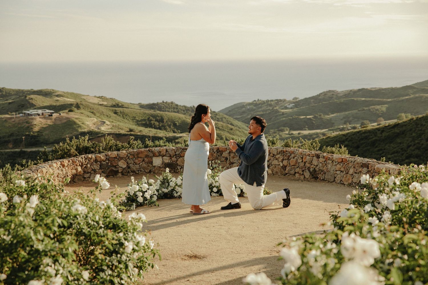 Man proposing to woman on scenic hilltop overlook with ocean views and white floral arrangements at sunset.