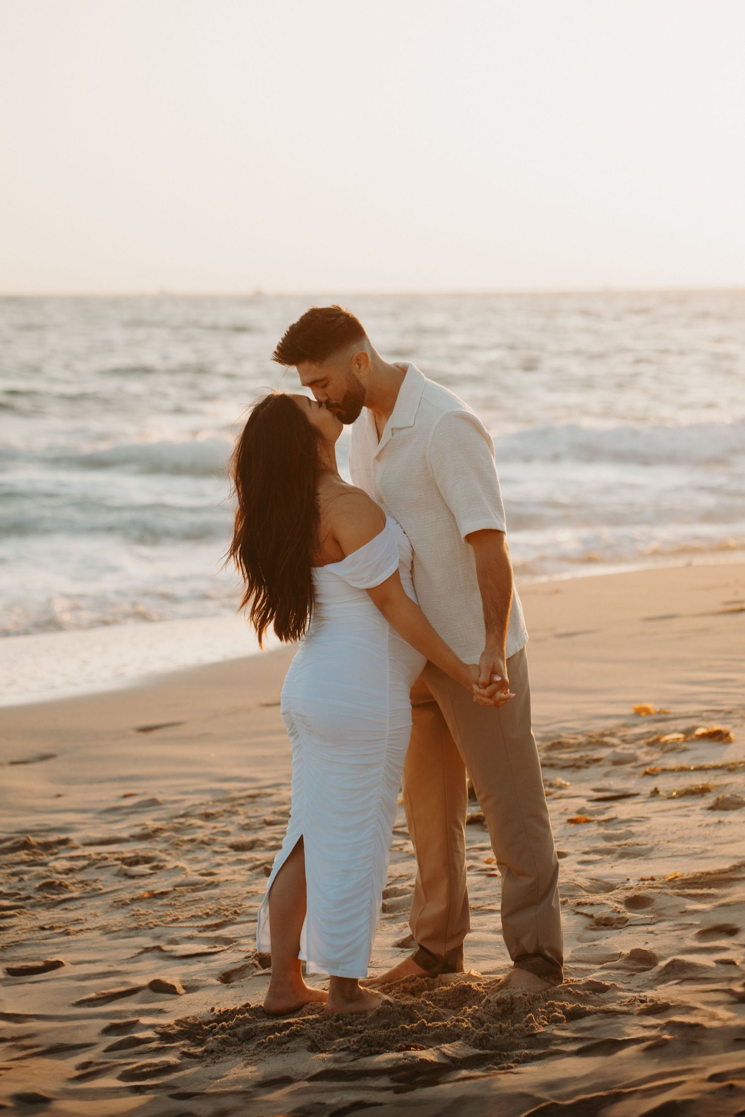 A romantic couple in white outfits shares intimate moments on a sandy beach at sunset.