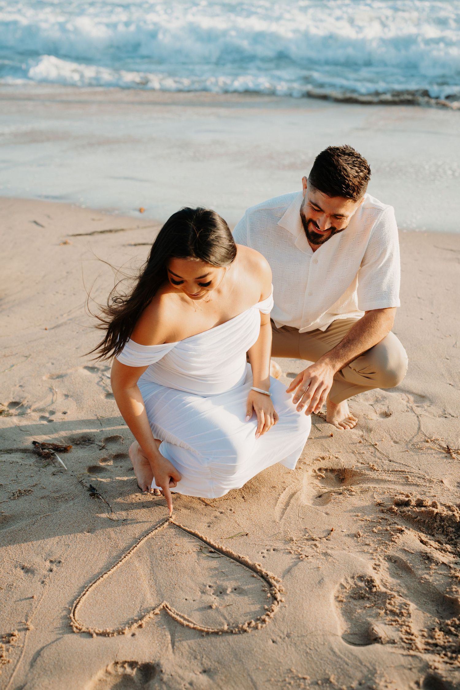 A couple draws a heart shape in the sand on a sunny beach during a romantic photoshoot.