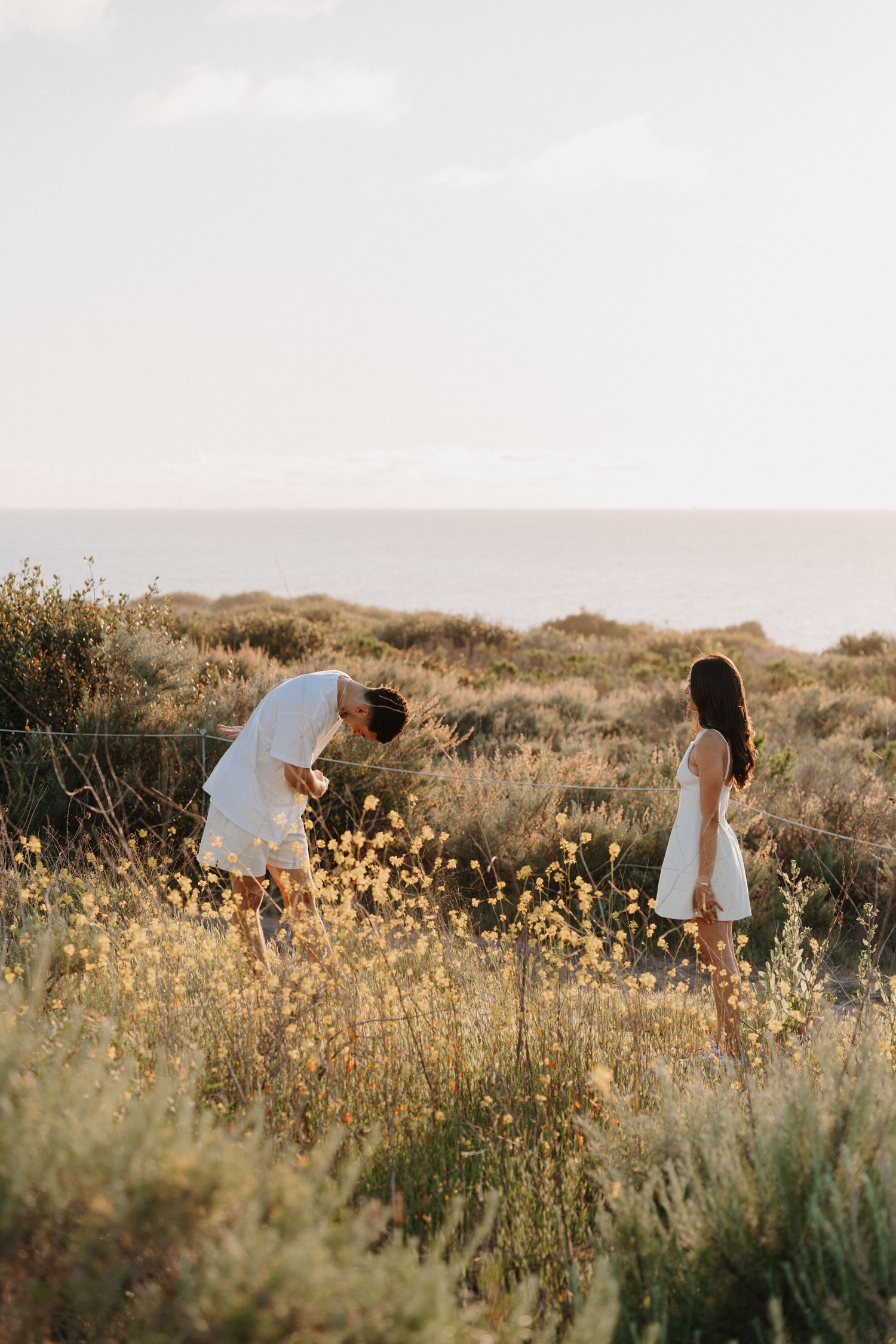 Two people in white outfits standing in a golden wildflower field at sunset near the ocean.