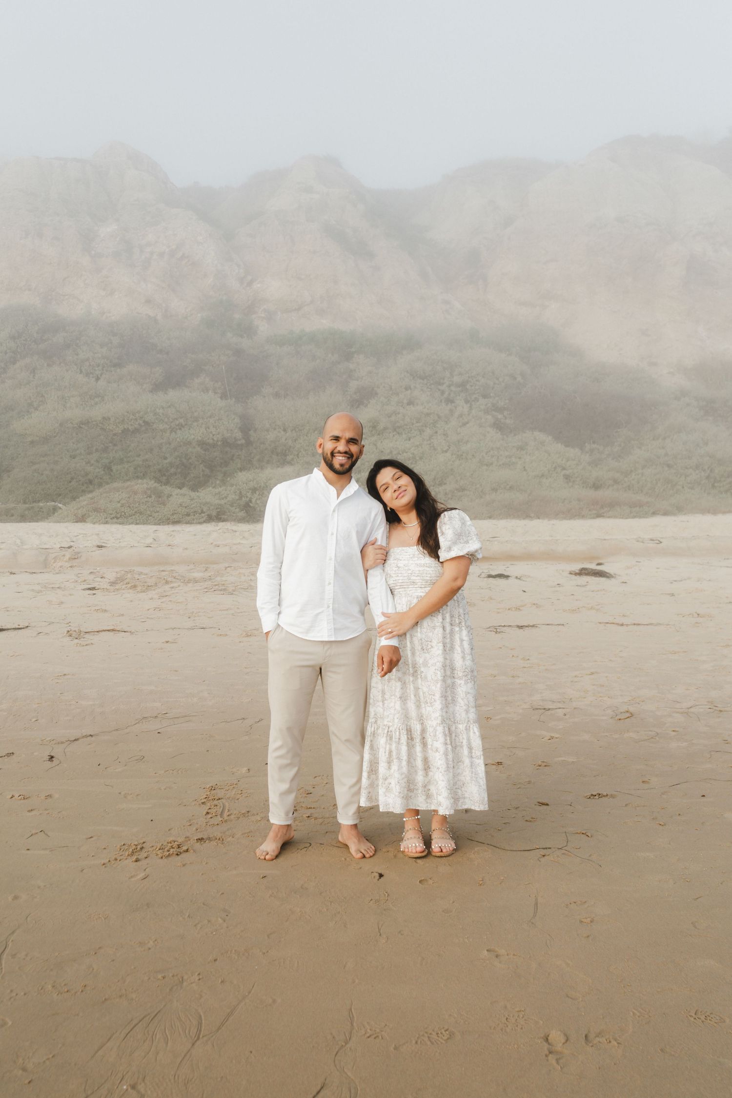A couple in white outfits poses together on a misty beach with mountains in the background.