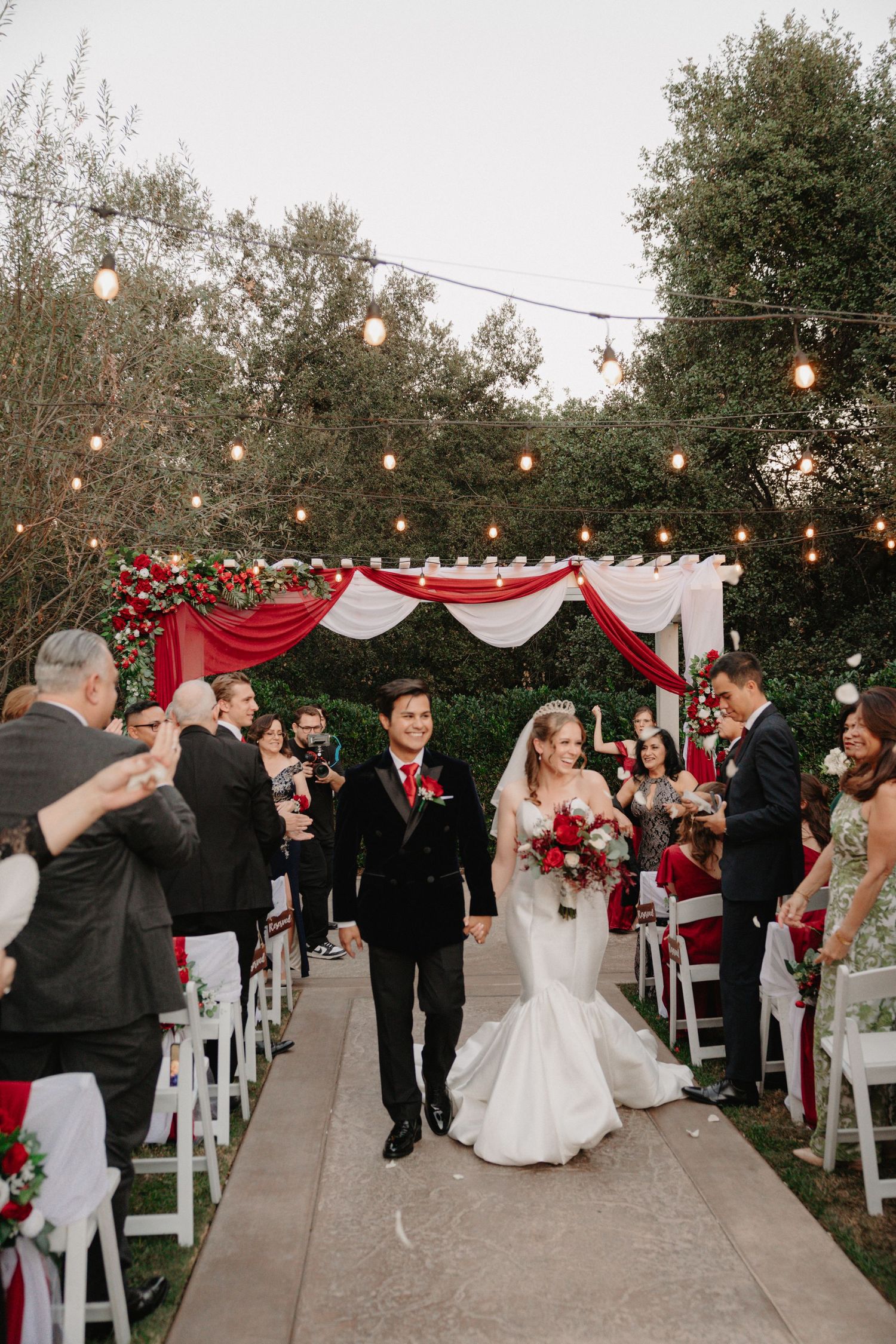 Outdoor wedding ceremony under string lights with red and white draping decor and guests celebrating.