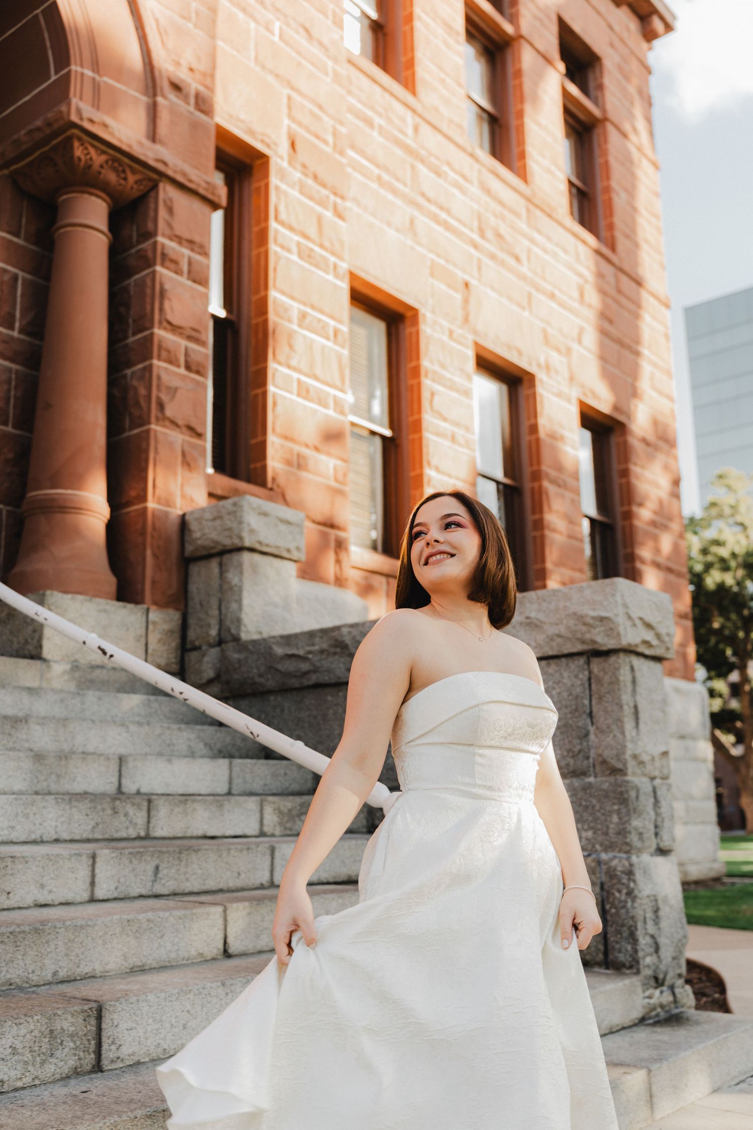 A bride in a strapless gown poses joyfully on stone steps near a brick building.