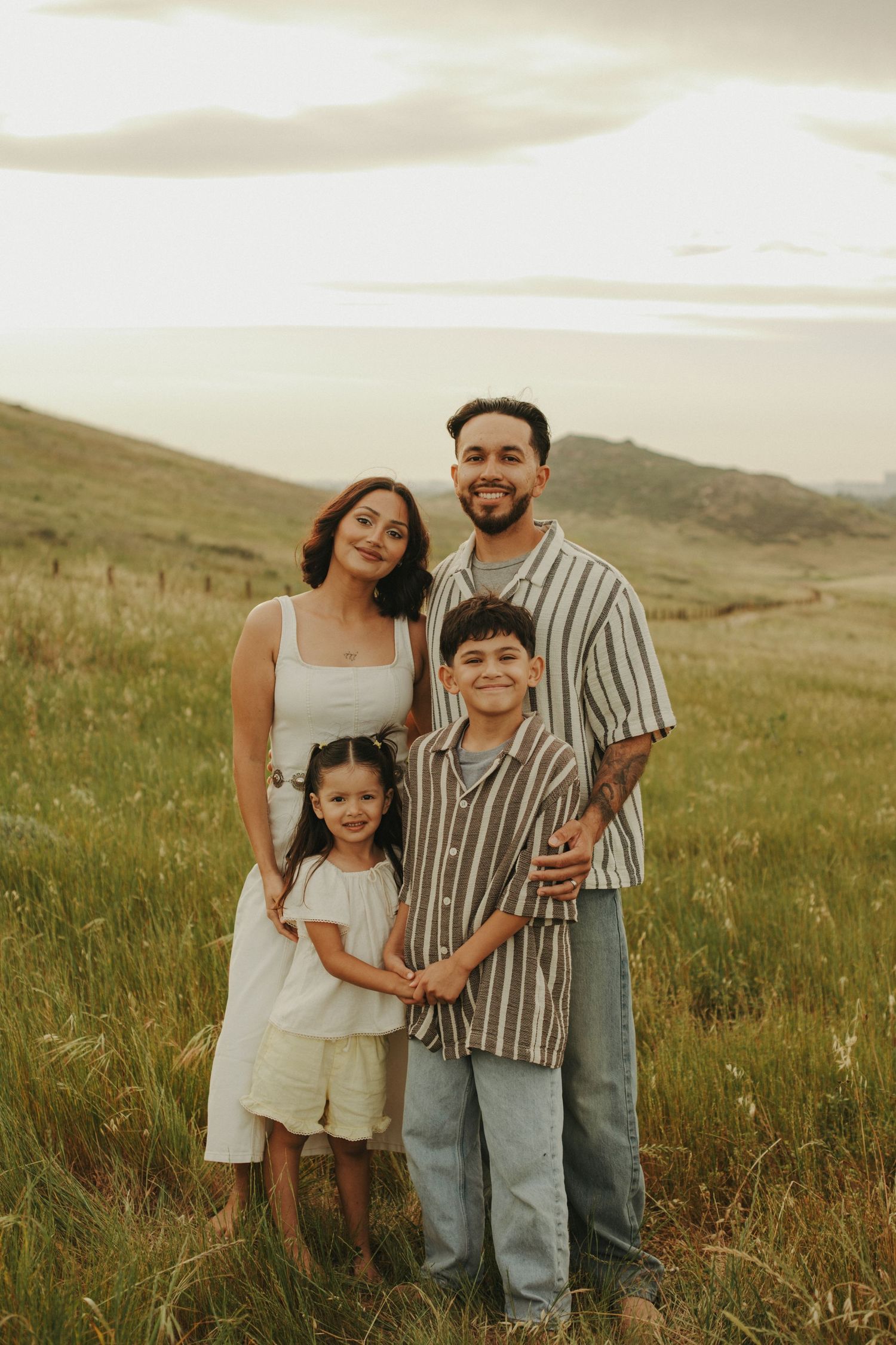 Family of four posing in a green meadow at sunset, parents and two children smiling in casual outfits.
