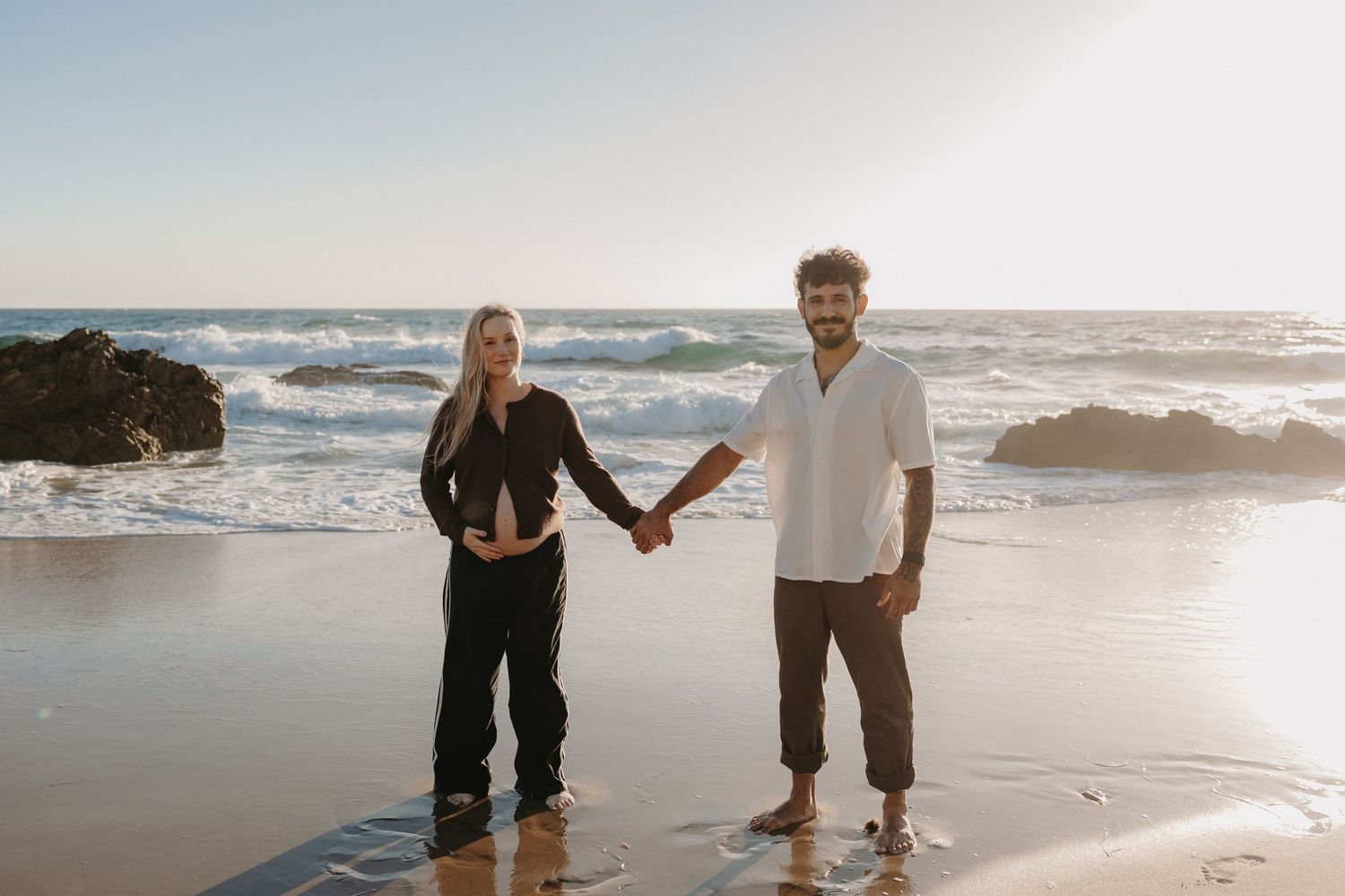 A couple walks together on a sandy beach at sunset with ocean waves crashing in the background.