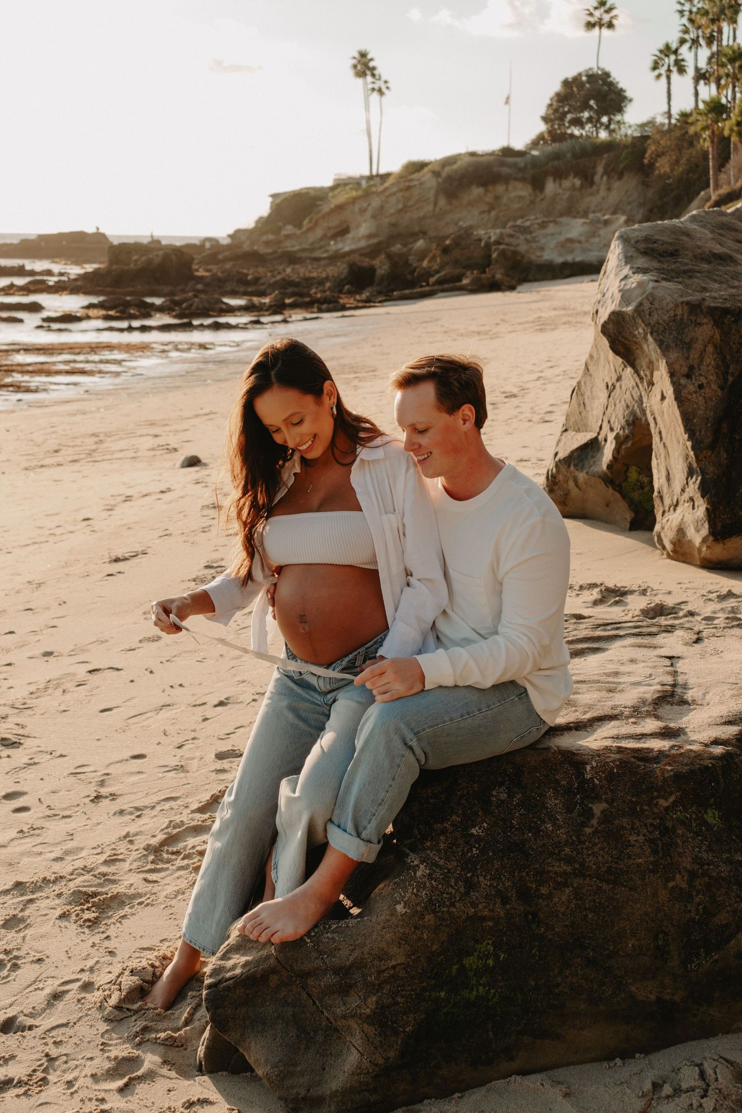 A couple shares intimate moments while sitting on rocks at a scenic beach during golden hour sunset.