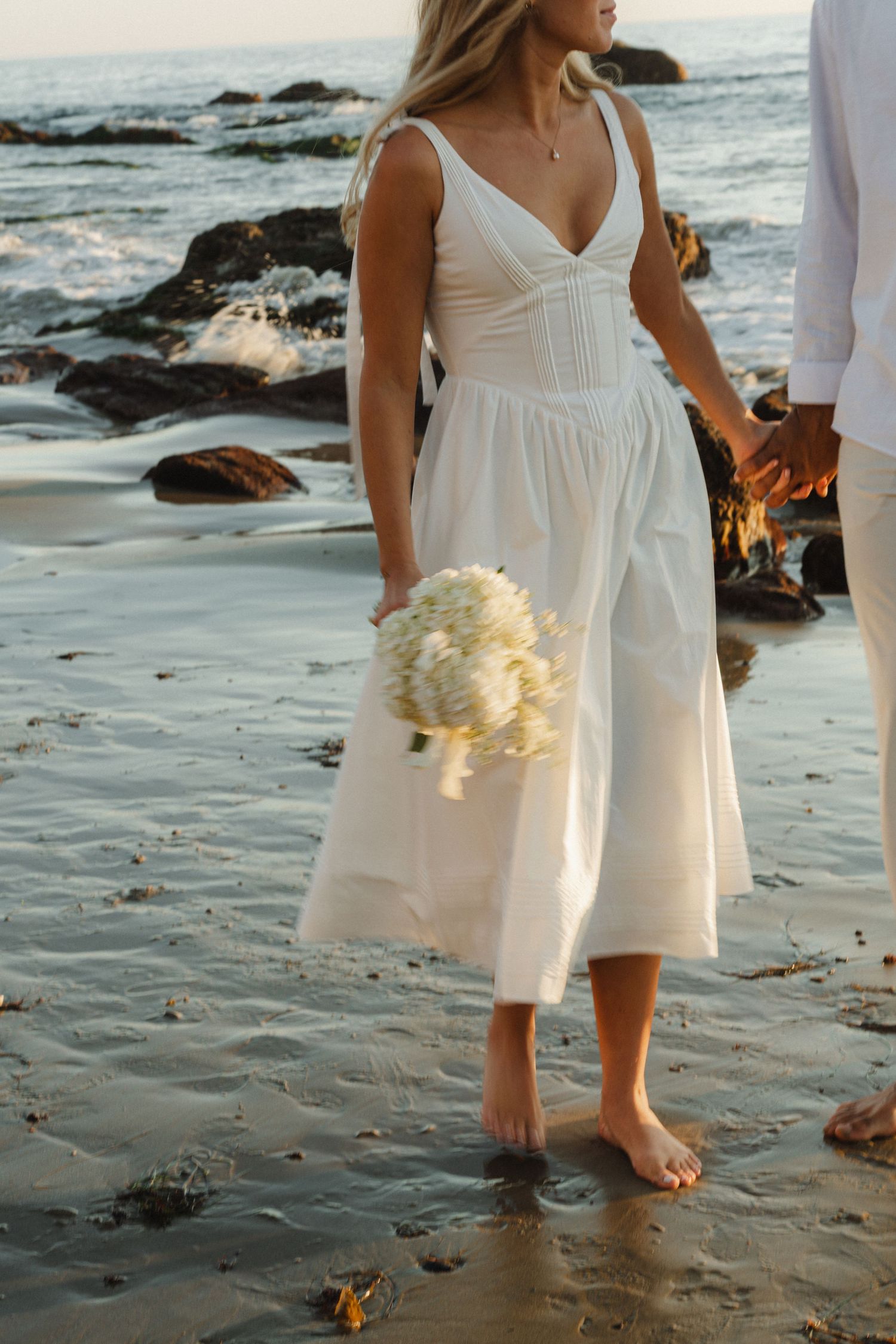 A person in a white flowy dress walks barefoot along a rocky beach at sunset.