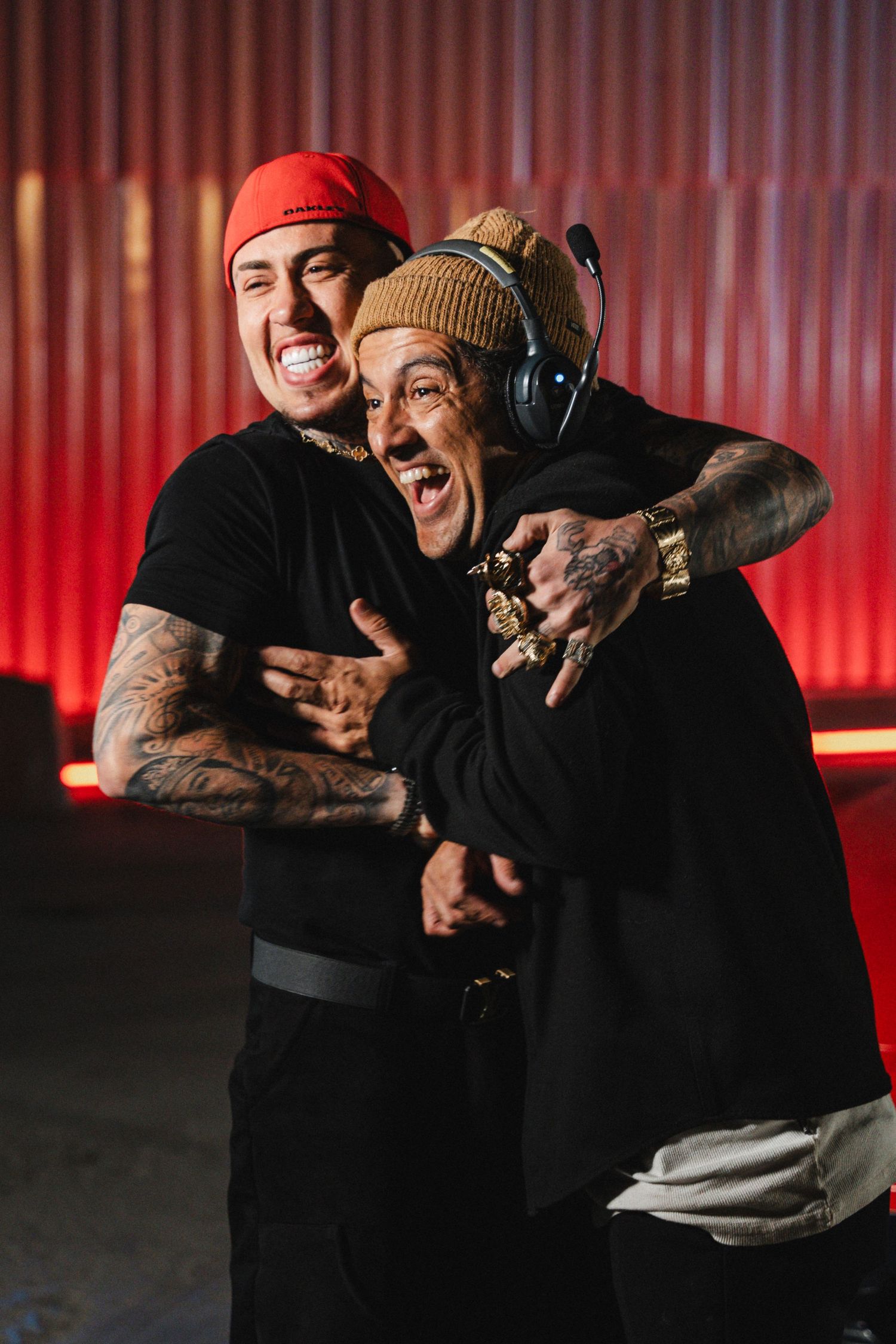 Two people in casual wear laughing and embracing in front of a red-lit backdrop.